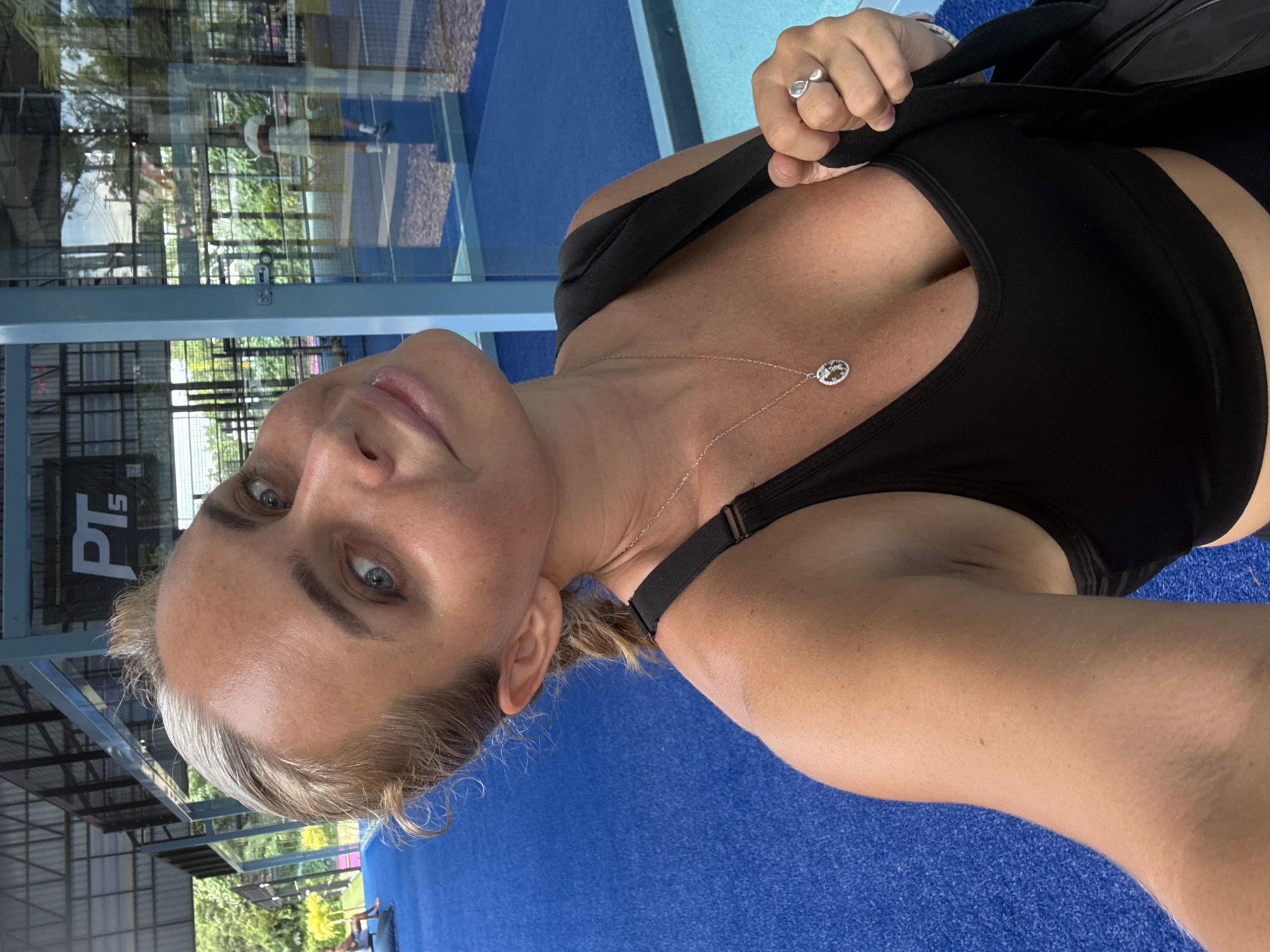 A woman is taking a selfie in an outdoor sports facility. She has blonde hair and blue eyes and is wearing a black sports bra and a silver necklace. She is holding a black bag over her shoulder and has a ring on her finger. The background shows a blue court with glass walls and some trees. The facility appears to be a padel court. The image is taken in the city of Ban Bang Rak, Thailand.