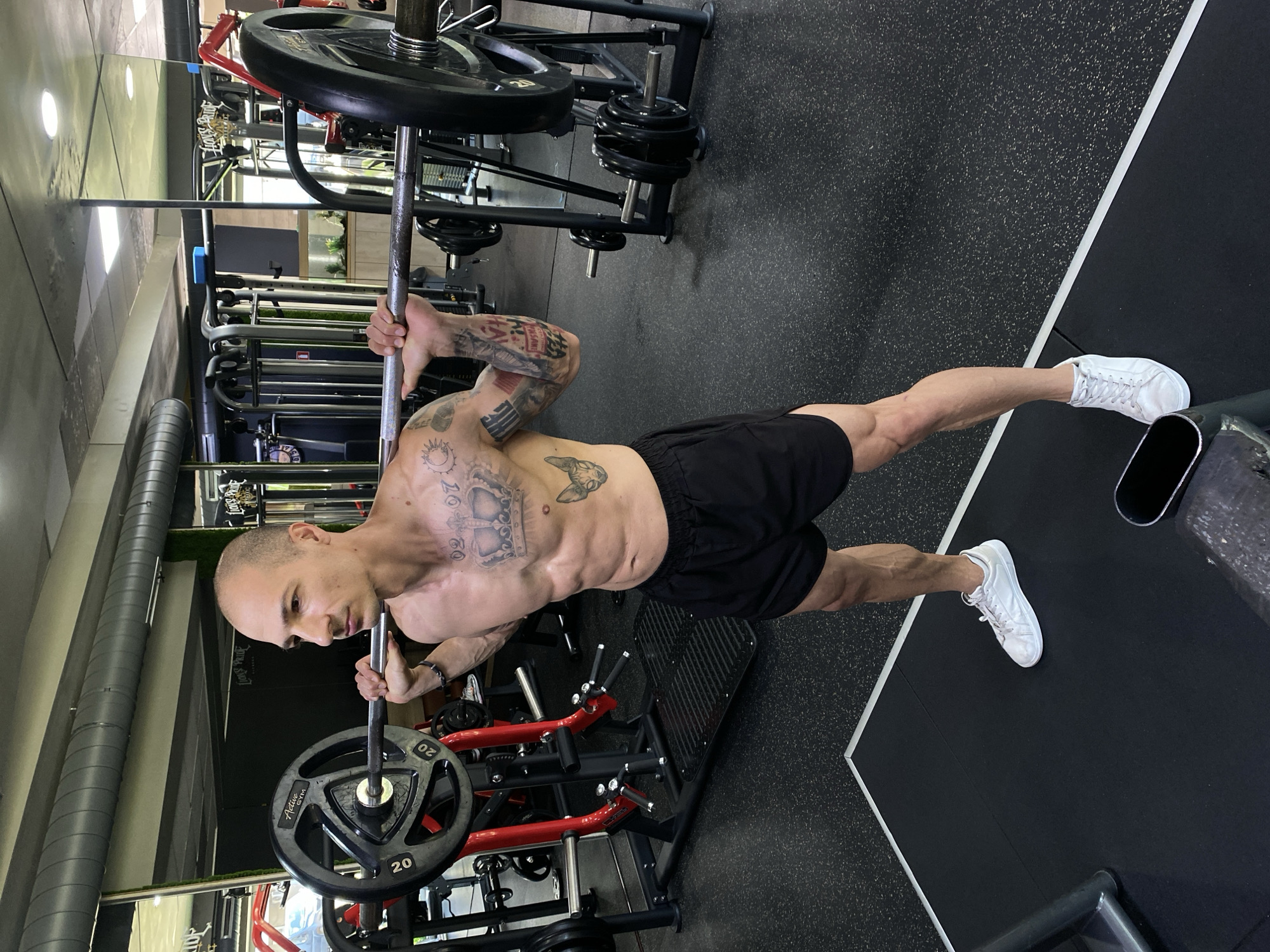 An athletic, bald man with a muscular physique is captured mid-workout in an indoor gym, located in Pleven, Bulgaria. He is shirtless, revealing extensive tattoos on his arms, chest, and torso, and wears black athletic shorts and white sneakers.
The man is performing an exercise, holding a barbell that appears to be anchored at one end as part of a T-bar row or landmine row machine, which features red framing and black platforms. A 20kg weight plate, marked with "20" and "Active Gym," is visible on the barbell. His right foot is placed on a raised black platform, while his left leg is extended straight backward, indicating a lunge or split-stance variation of the exercise. His gaze is directed downwards, suggesting intense focus and determination during the lift.
The gym environment features black rubber flooring with white dividing lines, various other workout machines and weight racks in the background, and large grey ventilation pipes running along the ceiling. The lighting is bright and even, typical of an indoor fitness facility, suggesting it is daytime.
Notable tattoos on his body include a crown with the date "10.08.80" on his left chest/ribs, a cat or sphinx-like figure on his right side, and numerous other symbols and script covering his arms. A blurry sign in the background might read "Lions Pride".
