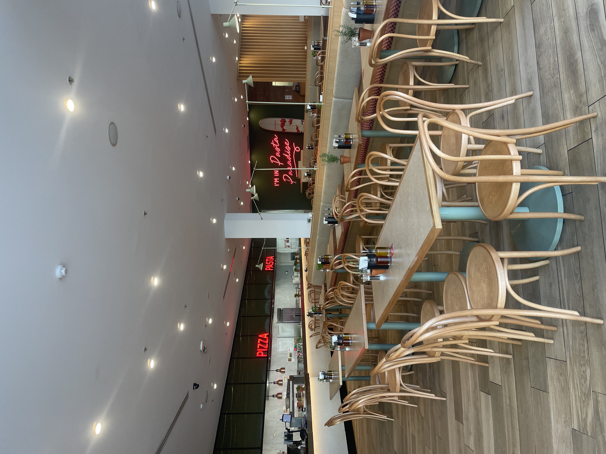 The image depicts the interior of a restaurant in Surfers Paradise, Australia. The restaurant has a modern and clean aesthetic with light wooden floors and tables. Red neon signs on the wall display the words "PIZZA" and "PASTA," indicating the restaurant's specialization. The seating arrangement includes several wooden chairs and banquette seating along the walls. The tables are set with condiments, including bottles of oil and vinegar. In the background, a counter area is visible with kitchen equipment and what appears to be a food preparation space. Another neon sign in the background reads "I'm in Pasta Paradise." The overall scene suggests a casual dining environment.