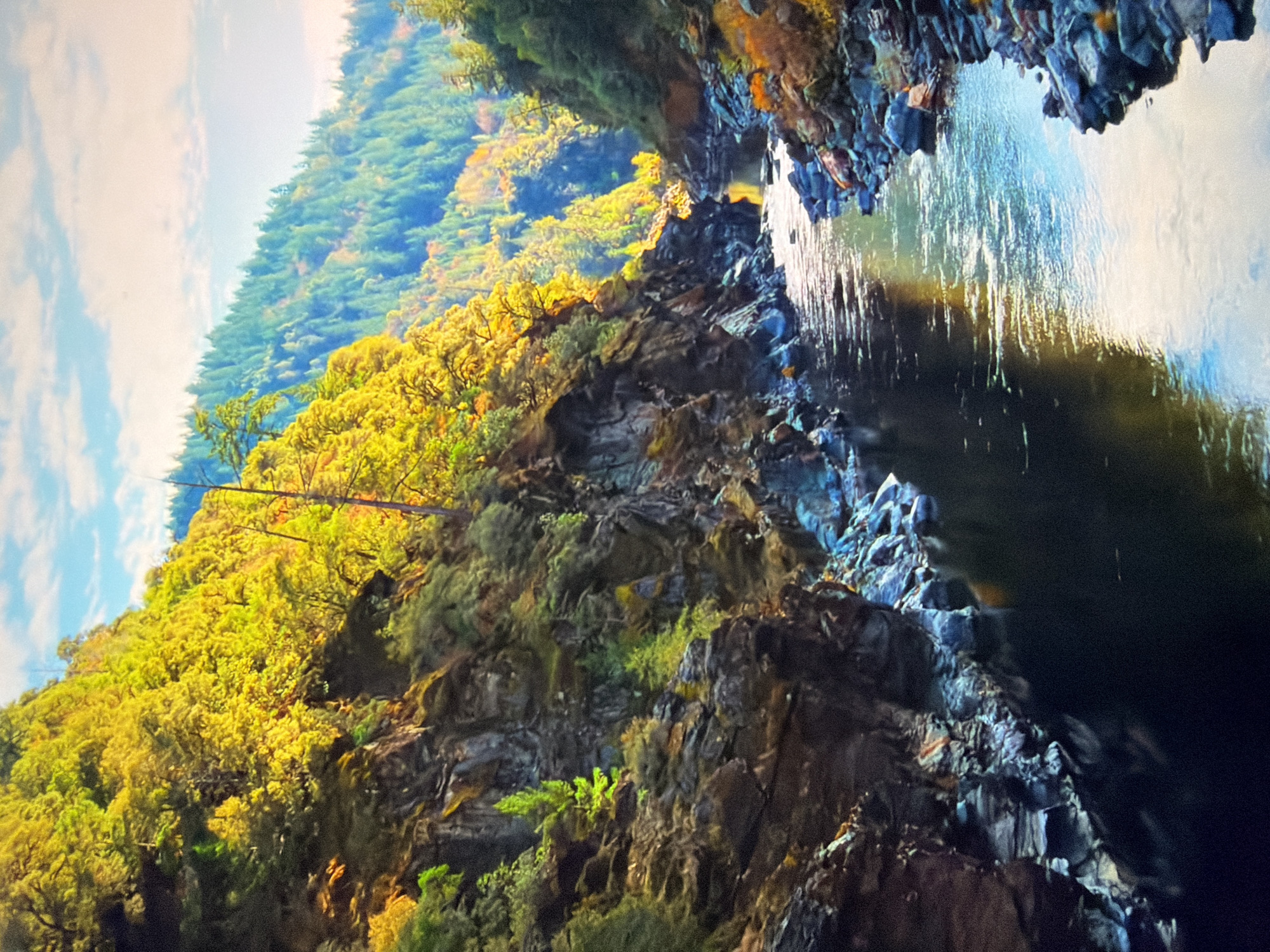 The image depicts a picturesque natural landscape. A wide river flows through a canyon with steep, rocky sides. The slopes are covered with dense vegetation, featuring trees in vibrant autumn foliage, with hues of yellow, green, and orange dominating the scene. The sky above is a clear blue with scattered white clouds. The water in the river is calm and reflects the surrounding landscape and sky. The overall impression is one of natural beauty and tranquility. The location is not specified as Kyiv, Ukraine.