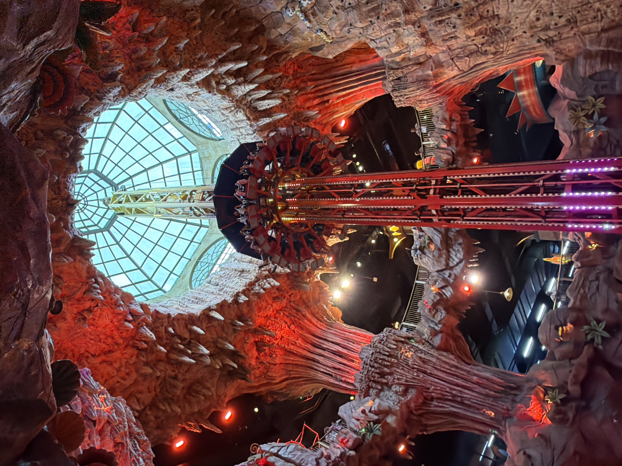The image shows the interior of a theme park, likely Sun World Ba Na Hills in Da Nang, Vietnam. A tall drop tower ride, with a circular passenger platform at the top, dominates the center of the frame. The ride structure is red and illuminated with purple lights. The surrounding environment is designed to resemble a cave or a rocky, prehistoric landscape, with large, textured rock formations and stalactites. The rock surfaces are illuminated with red and orange lights, creating a dramatic and fiery ambiance. A large, domed glass ceiling with a grid pattern is visible at the top of the structure, allowing natural light to filter in, casting a blue-green hue. Several people are visible on the ride platform and on walkways below. The overall scene depicts an indoor amusement attraction with a fantasy or adventure theme.