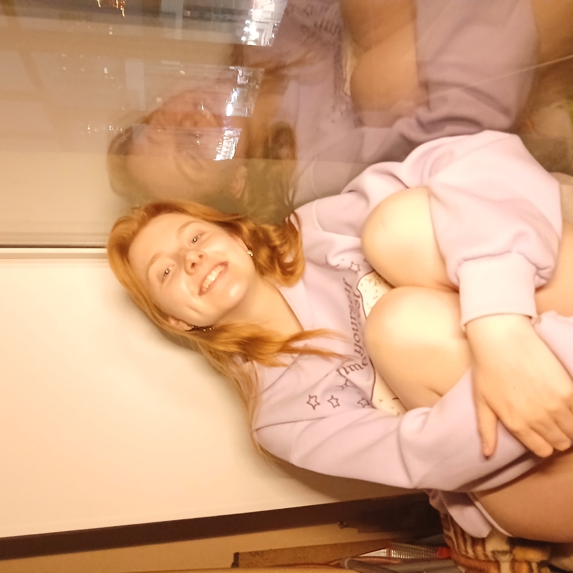 An indoor, medium close-up portrait features a young female with long, reddish-blonde hair. She is lying or sitting on her side, with her knees drawn up towards her chest, and is looking directly at the camera with a wide, open-mouthed smile, showing visible teeth. Her expression conveys happiness and comfort.
She is wearing a light purple long-sleeved top or hoodie, which features white star motifs and partially visible cursive text on its side. The words "yourself" and "me" can be discerned on the fabric. A small portion of a white undergarment is visible near her left leg.
The background primarily consists of a plain, light-colored wall or surface. A prominent feature is a large reflective surface, likely a window or glass panel, that occupies the upper and left sections of the image. This surface reflects a blurry, inverted image of the subject's upper body and hair, as well as abstract, out-of-focus lights, suggesting an urban exterior or another brightly lit interior beyond the glass. The soft, warm lighting suggests an evening or nighttime setting.
The overall scene is intimate and personal. While specific geographical cues are not visible within the image, the provided context places the location in Yekaterinburg, Russia.