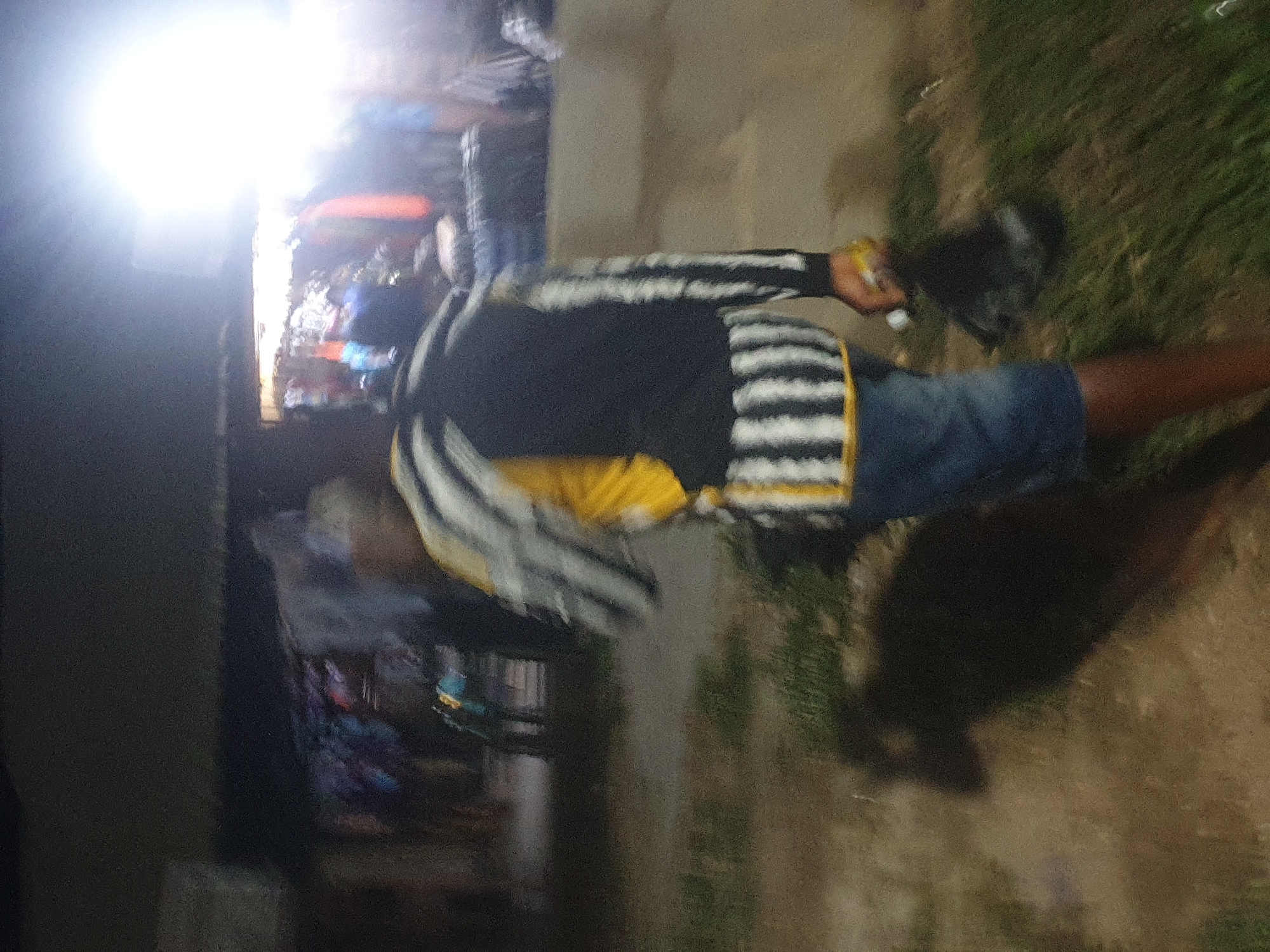 The image shows a person outdoors in Zango-ndaji, Nigeria. The person is wearing a black and white striped long-sleeved shirt, a yellow shirt underneath, denim shorts, and is holding a dark-colored object in their hand. The person is standing on a dirt or gravel surface next to some grass. Behind the person are buildings, and there appears to be a bright light source visible.