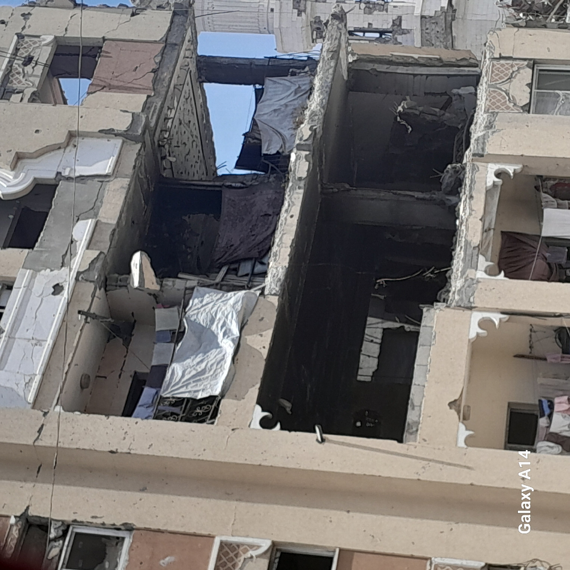 This image depicts the severe damage to a multi-story building in Khan Yunis, Palestinian Territories, characterized by extensive structural damage, debris, and visible signs of conflict. The facade shows large sections of the exterior wall crumbling, exposing inner structural elements. Several windows are either broken or entirely missing, revealing dark, debris-filled interiors. Laundry, appearing to be white and colored fabrics, is draped over some of the remaining balconies and structures, suggesting ongoing or recent habitation despite the destruction. The overall scene is one of devastation, with clear evidence of the building having sustained significant impact, likely from military action. A minaret of a mosque is partially visible in the background to the right, indicating a densely populated urban environment. The sky is clear and blue, contrasting with the destroyed architecture. The lower left corner of the image contains a watermark identifying the device as "Galaxy A14".