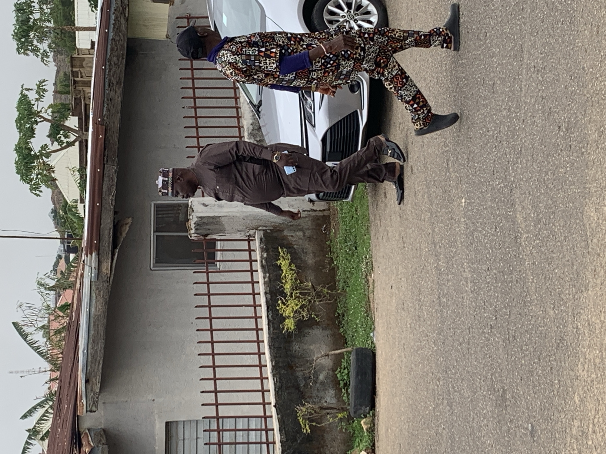 Two adult males are walking along a paved road in what appears to be a residential area of Jos, Nigeria. The man closest to the camera is wearing a brown suit with a traditional cap. He is holding a light-colored object, possibly a phone. The other man is wearing a patterned suit with a black cap. Both are wearing dark shoes. A white car is parked behind them. A building with a rusty metal roof, a window, and a dark brown metal fence stands to their left, and a patch of greenery is near the fence.