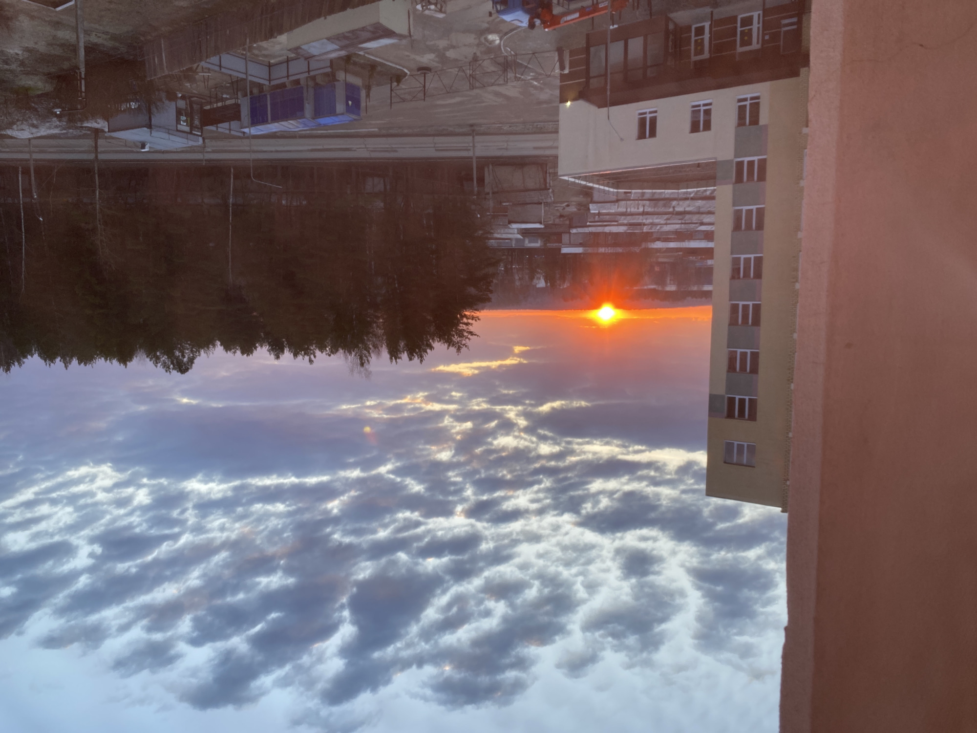 The image, when correctly oriented, presents a vibrant sunset or sunrise viewed from a high vantage point, likely a building's window or balcony, overlooking Chaikovsky, Russia.
The scene is dominated by a striking sky and a mixed landscape. In the immediate foreground on the right, a warm, reddish-brown wall or balcony railing partially frames the shot. To the left, a multi-story residential building with light-colored (beige and grey) facades and numerous windows stands.
Beyond this building, a dense, dark forest stretches across the midground, its trees silhouetted against the bright horizon. Below the distant horizon and bordering the forest, industrial or construction elements are visible, including concrete surfaces, fencing, and some blue containers, suggesting an area of development or an industrial zone. A road or path runs alongside the edge of the forest.
The sun, a brilliant orange-red orb, is positioned low on the horizon, just above the distant tree line. The sky is a spectacular blend of colors, transitioning from intense oranges and yellows near the sun to softer pinks, purples, and blues higher up. It is filled with a variety of clouds; some are dark and undulating, while others catch the warm glow of the sun, creating intricate patterns and highlights.
No people are visible. The overall impression is one of natural beauty, specifically a colorful atmospheric event, set against a backdrop that combines dense woodland with urban and industrial structures. The time of day is clearly sunset or sunrise due to the low angle and intense color of the sun. There is no visible text in the image.