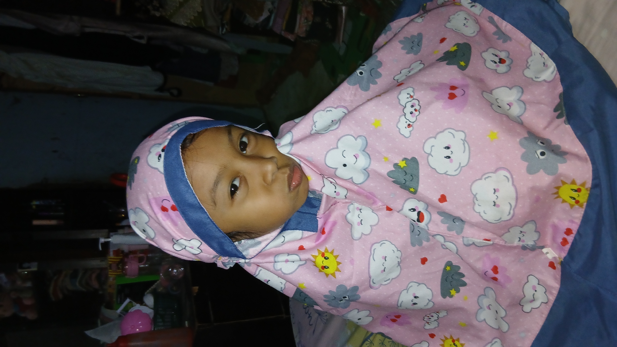 In Keudejungkagajah, Indonesia, a young girl, appearing to be around 4-6 years old, is the central subject of the image. She is wearing a pink hijab and matching fabric adorned with a pattern of cartoonish clouds, suns, and hearts. The girl is looking directly at the camera with a pout. She is situated in what appears to be a dimly lit indoor environment, possibly a bedroom or living area. The background is cluttered with shelves containing various items, including what look like stuffed animals, bottles, and other household objects. The lighting suggests it is nighttime or a poorly lit indoor space.