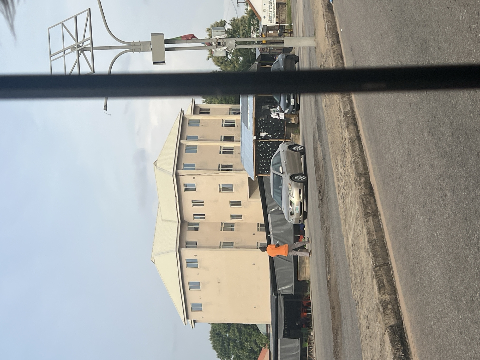 The image depicts a street scene in Jalingo, Nigeria. In the background, a three-story building with a cream-colored facade and a light-colored roof dominates the view. The building has numerous windows distributed across its levels. In front of the building, there is a covered structure that appears to be a shop or outdoor seating area, with black fabric covering its sides and a blue corrugated metal roof.
On the street, a man wearing an orange t-shirt and grey pants is walking from left to right. To his right, a silver sedan is parked or moving slowly. Further to the right, another dark-colored sedan is visible, and a person in a white shirt and dark pants is standing near the covered structure. On the right side of the image, a lamppost with a solar panel array is prominent, and a flag is attached to it. Signs are also visible on buildings in the background on the right.
The foreground shows a section of a paved road with a raised concrete curb. The road surface appears worn, with some patches of exposed earth and gravel along the curb. The sky above is a pale blue with some cloud cover. The overall impression is of a typical urban environment with commercial activity.