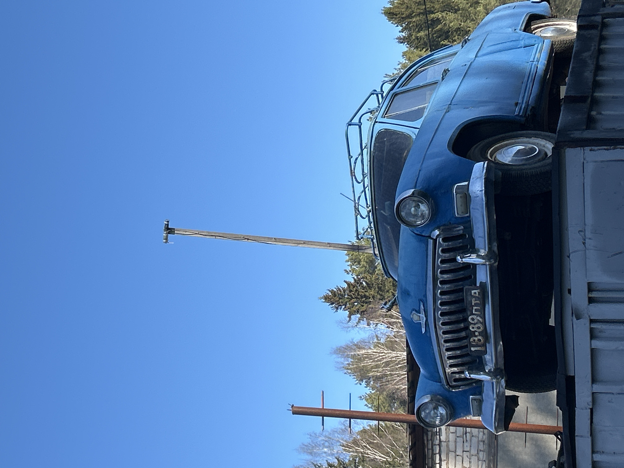 The image shows a blue vintage car on a flatbed truck or trailer. The car has a roof rack. The license plate is visible and reads "18-89 ПТА." The background includes a clear blue sky, trees (some with snow on the ground), and a utility pole. Based on the license plate, the location is likely in Russia, specifically the city of Chaikovsky.
