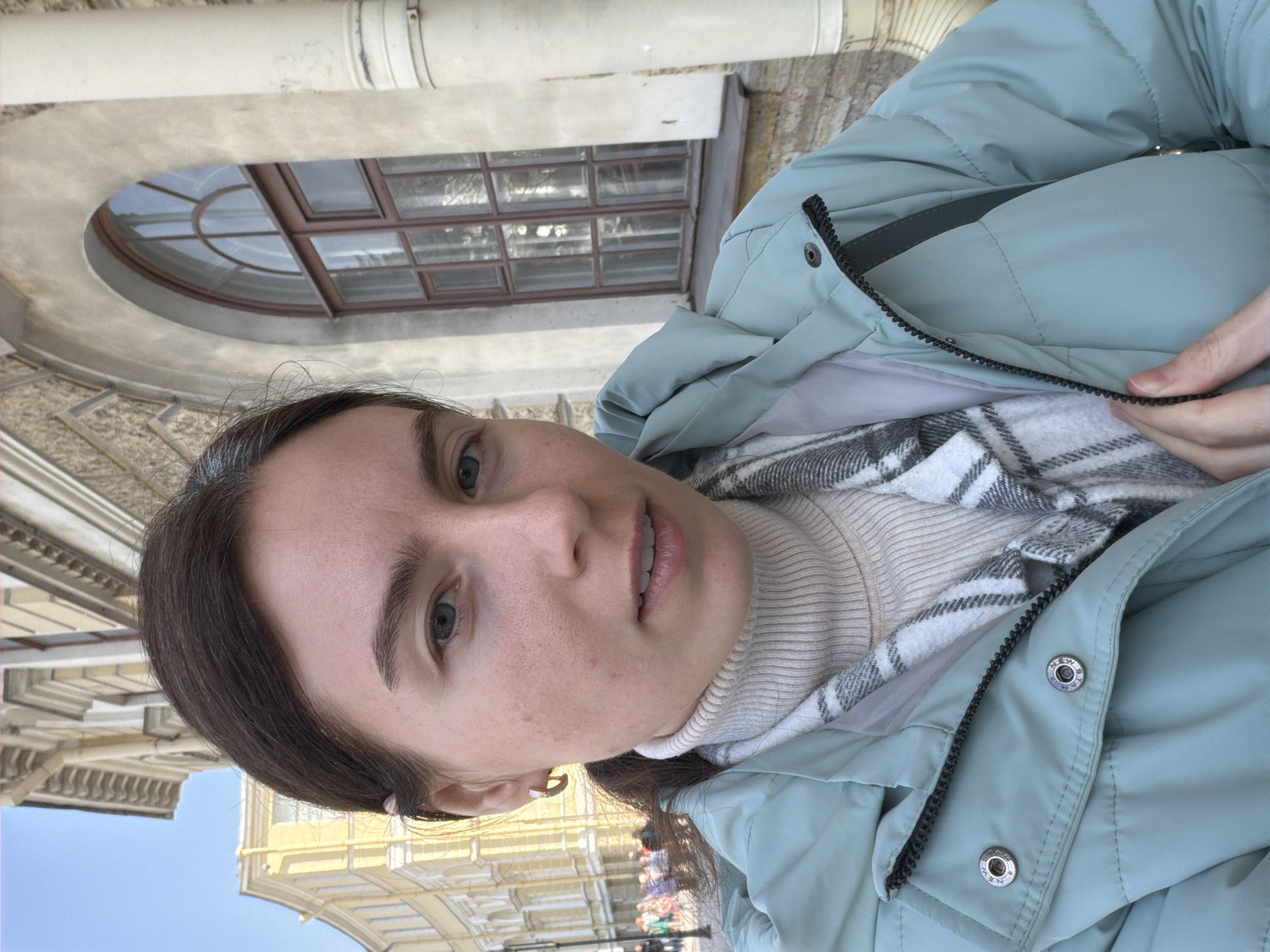 A woman wearing a light blue puffer jacket, a beige turtleneck, and a gray and white checkered scarf is taking a selfie outdoors. The background shows architectural details of Saint Petersburg, Russia, including an arched window with a grid pattern, decorative molding, and structures suggesting a street scene. The sky is partly visible in the upper right corner. A group of people is blurred in the background.