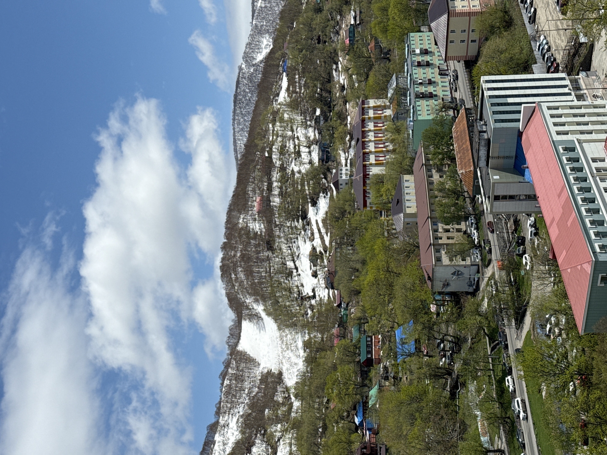 This bright, sunny daytime image presents an expansive view of a city nestled against the base of a large, snow-capped mountain range, captured from an elevated perspective. The sky is a clear blue, adorned with scattered white, puffy clouds.
The dominant feature in the mid-ground is a mountain whose slopes are covered in a mix of bare trees and evergreen foliage, with significant patches of snow still visible, particularly in shaded areas and higher elevations. This suggests a late spring or early summer setting.
In the foreground and lower mid-ground, the city unfolds with a dense arrangement of multi-story buildings. These structures display a variety of architectural styles and colors, including light green, yellow, beige, and white facades, many featuring prominent red or brown roofs. Some buildings appear to be residential apartment blocks, while others could be commercial or public facilities.
Numerous green trees with fresh spring leaves are interspersed throughout the urban landscape, lining streets and dotting courtyards. Paved roads wind through the city, with many cars visible, both parked along curbsides and in designated parking areas. The scene depicts typical urban life, with no specific human activity or identifiable individuals highlighted.
The overall impression is one of a vibrant city in a dramatic natural setting. Based on the provided context, the location is Petropavlovsk-Kamchatsky, Russia, known for its mountainous terrain and coastal environment. No legible text is visible on any buildings or signs within the image.