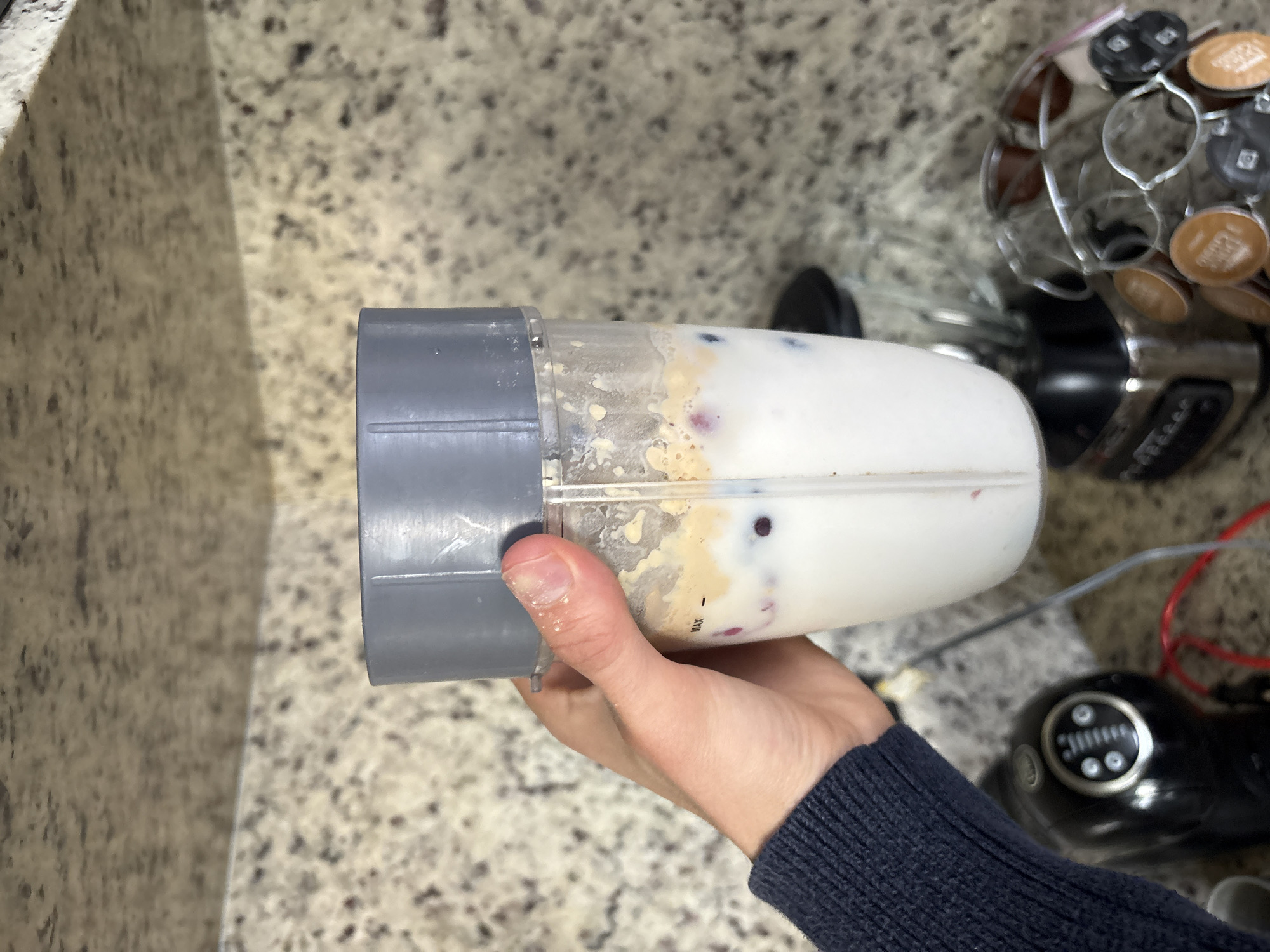A person holds a blender cup filled with a smoothie. The smoothie appears to contain blueberries. The background shows a countertop with speckled granite, a Nespresso machine and a coffee pod holder. The location is not discernible from the image.