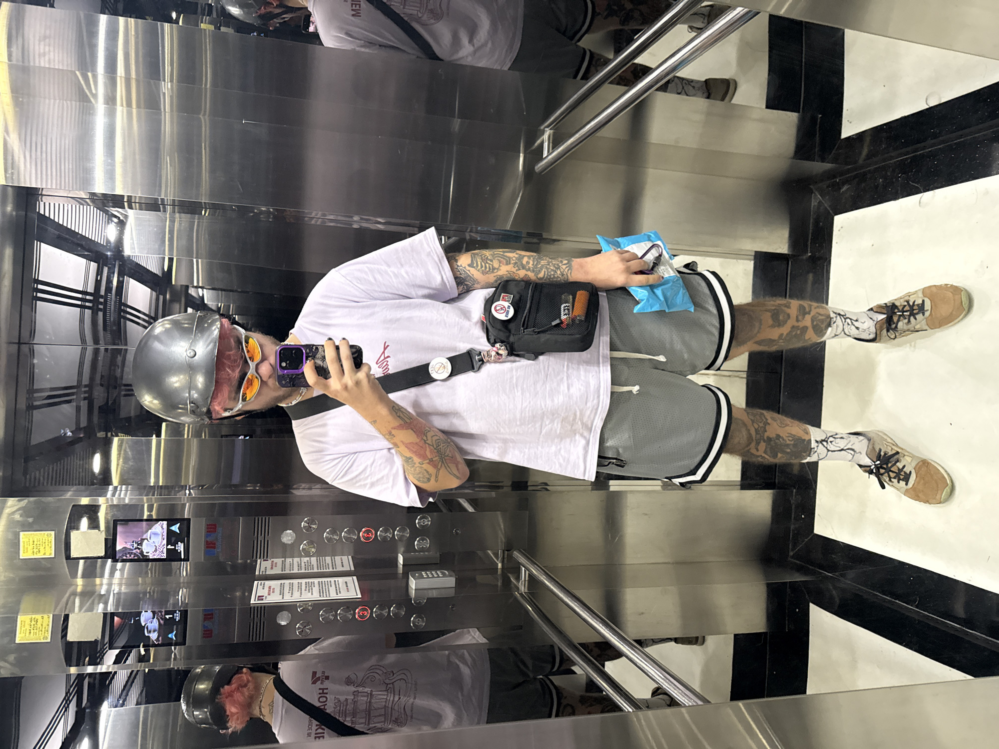 The image depicts a person taking a selfie in an elevator. The person is wearing a silver helmet, orange-tinted sunglasses, a white t-shirt, gray athletic shorts, and patterned socks with tan shoes. They are holding a phone in their right hand and a small blue package in their left. The person has tattoos on their arms and legs. They are also wearing a small black bag across their chest.
The elevator has a mirrored interior, stainless steel panels with control buttons, and a digital display showing the floor number. The floor of the elevator is tiled with a black and white geometric pattern.
The location is likely in a building within the city of Đà Nẵng, Vietnam.