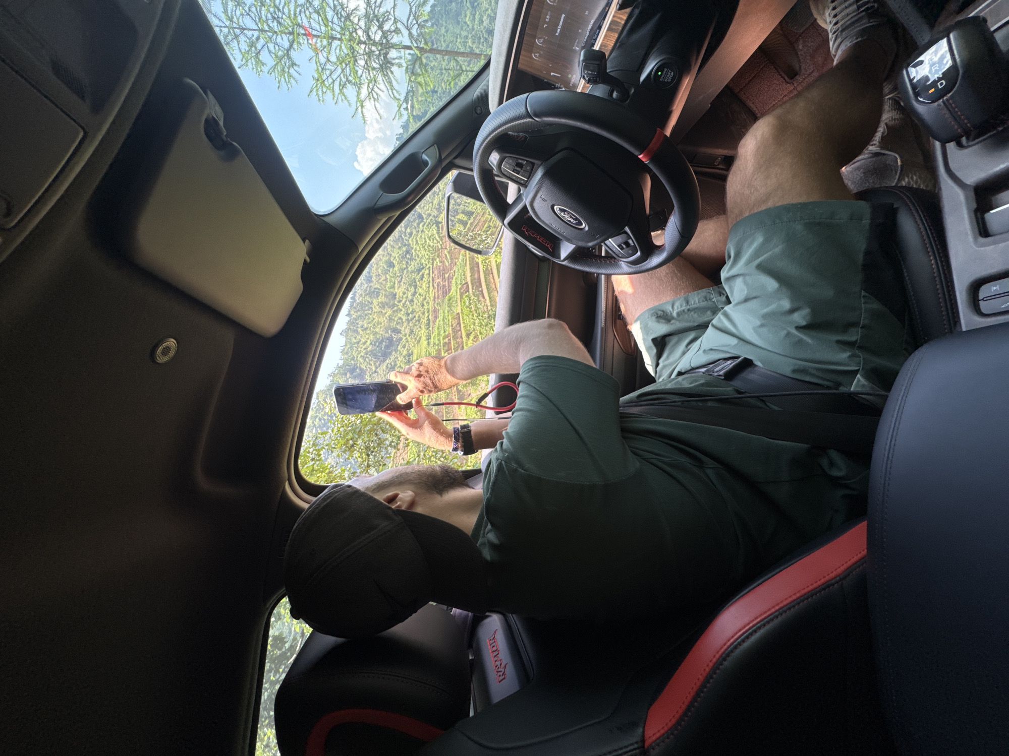 A man in a black cap and green shorts is seated in the driver's seat of a Ford truck. He is wearing a black watch on his left wrist and holding a cell phone with both hands to take a picture of the landscape seen through the driver's side window. The car's interior includes a black and red stitched seat and a Ford steering wheel. Outside, the landscape shows green vegetation and a clear sky, consistent with a mountainous or rural setting. This image was taken in Vinh Quang, Vietnam.