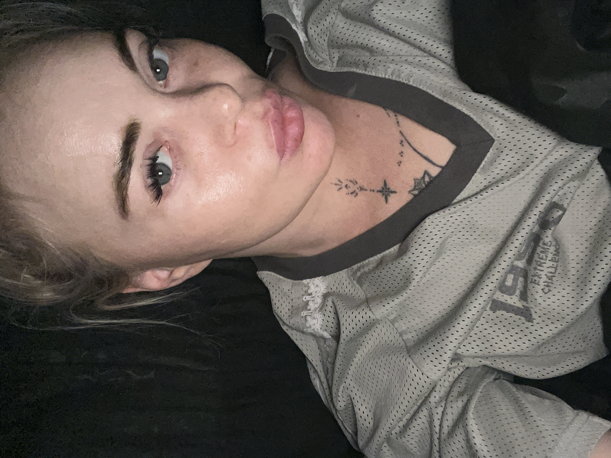 A close-up, selfie-style photo shows a light-skinned woman with blonde hair and blue eyes. She is wearing a light gray, mesh jersey with dark gray trim. The jersey has some white lace detailing on the collar. The woman has a tattoo on her neck that appears to be a star with some smaller markings around it. Her lips are slightly pursed, and she is looking directly at the camera. The background is dark and out of focus, suggesting an indoor setting with low lighting. The lower part of the jersey has text that is partially visible, appearing to say "1999 EXTREME CHALLENGE."