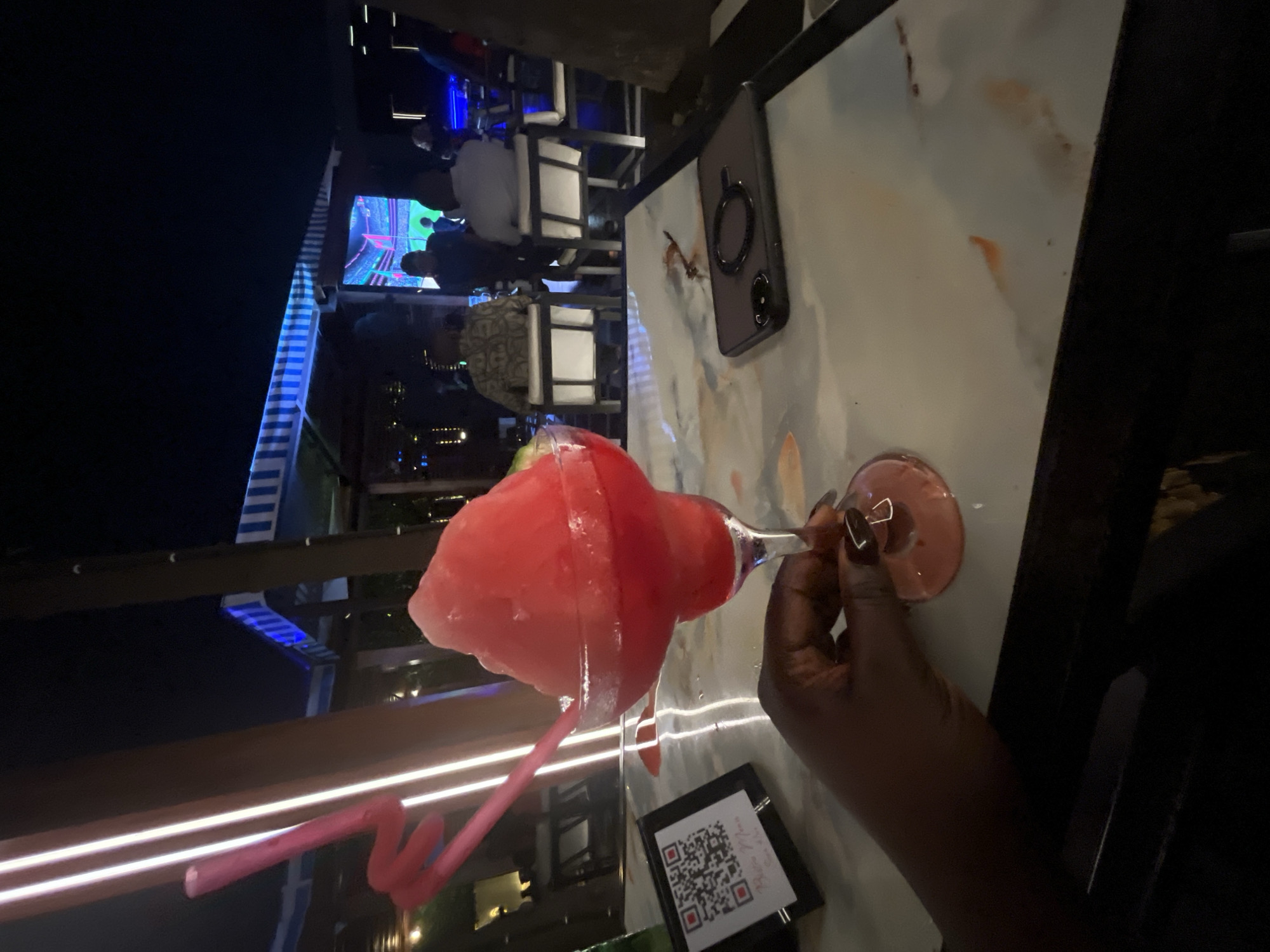 In the city of Abuja, Nigeria, at night, a person's hand with dark skin and manicured nails holds a large, frosty margarita glass filled with a pink, blended beverage. The glass has a pink, swirled straw inserted into it. The drink appears to be a strawberry or watermelon flavor due to its color. The beverage sits on a white marble-patterned table, which has a few small reddish-brown stains on it. To the left of the drink, a small black frame holds a QR code. A dark-colored smartphone with a black circular ring holder is placed on the table to the right of the drink. In the background, people are seated at tables in a dimly lit, outdoor setting. There are lights visible, including vertical white LED strips on the left, and a large screen displaying graphics in the center-right. The overall atmosphere suggests a bar or restaurant environment.
