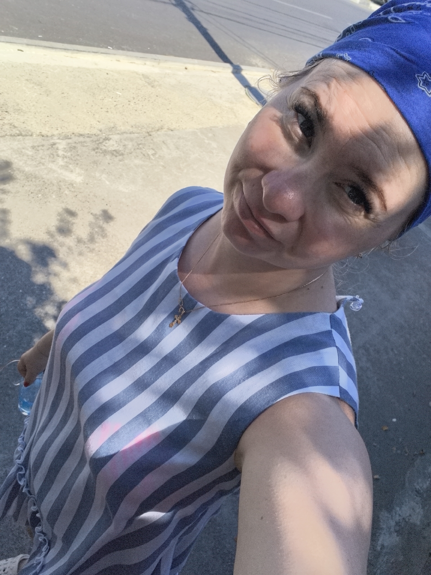 A woman in a blue and white striped dress and a blue bandana is taking a selfie outdoors in Vityazevo, Russia. The woman is looking at the camera with a slight smile. She is wearing a gold cross necklace. The background shows a paved area, possibly a sidewalk or road, with shadows cast from overhead objects. The lighting suggests it is daytime. The woman is holding a clear plastic bottle in her left hand.