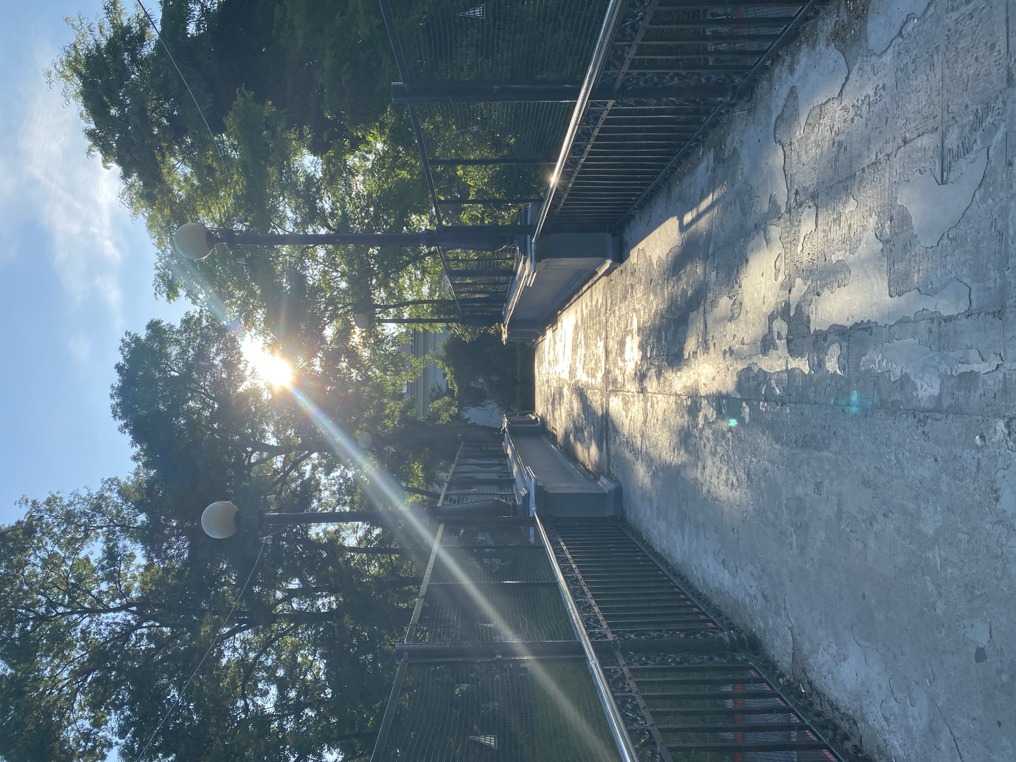 The image shows a pedestrian bridge at the city of Sochi, Russia, under a bright sun. The sun's rays are shining through the trees, creating lens flares and casting shadows on the bridge's walkway. The bridge has decorative metal railings and lampposts. The walkway appears to be made of concrete and shows signs of wear and tear, with patches of exposed material and some wet areas reflecting the sunlight. The background is filled with lush green trees and a clear blue sky.