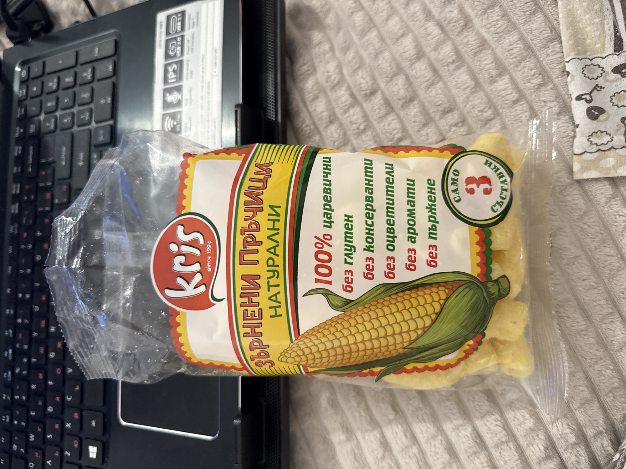 A close-up shot features a bag of "Kris" brand corn puffs resting partially on a black laptop and a textured, possibly quilted, surface. The bag has a clear plastic front, allowing visibility of the yellow corn puffs inside. The packaging is decorated with a cartoon illustration of an ear of corn. Text on the bag, in Bulgarian, highlights that the puffs are "100% corn," "natural," and free of gluten, preservatives, coloring agents, and artificial flavors. A small green circle indicates "3 ingredients." The laptop's keyboard is visible on the left side of the frame, and the texture of the surface is visible to the right of the bag. There's no evidence to suggest this is in Blagoevgrad, Bulgaria.