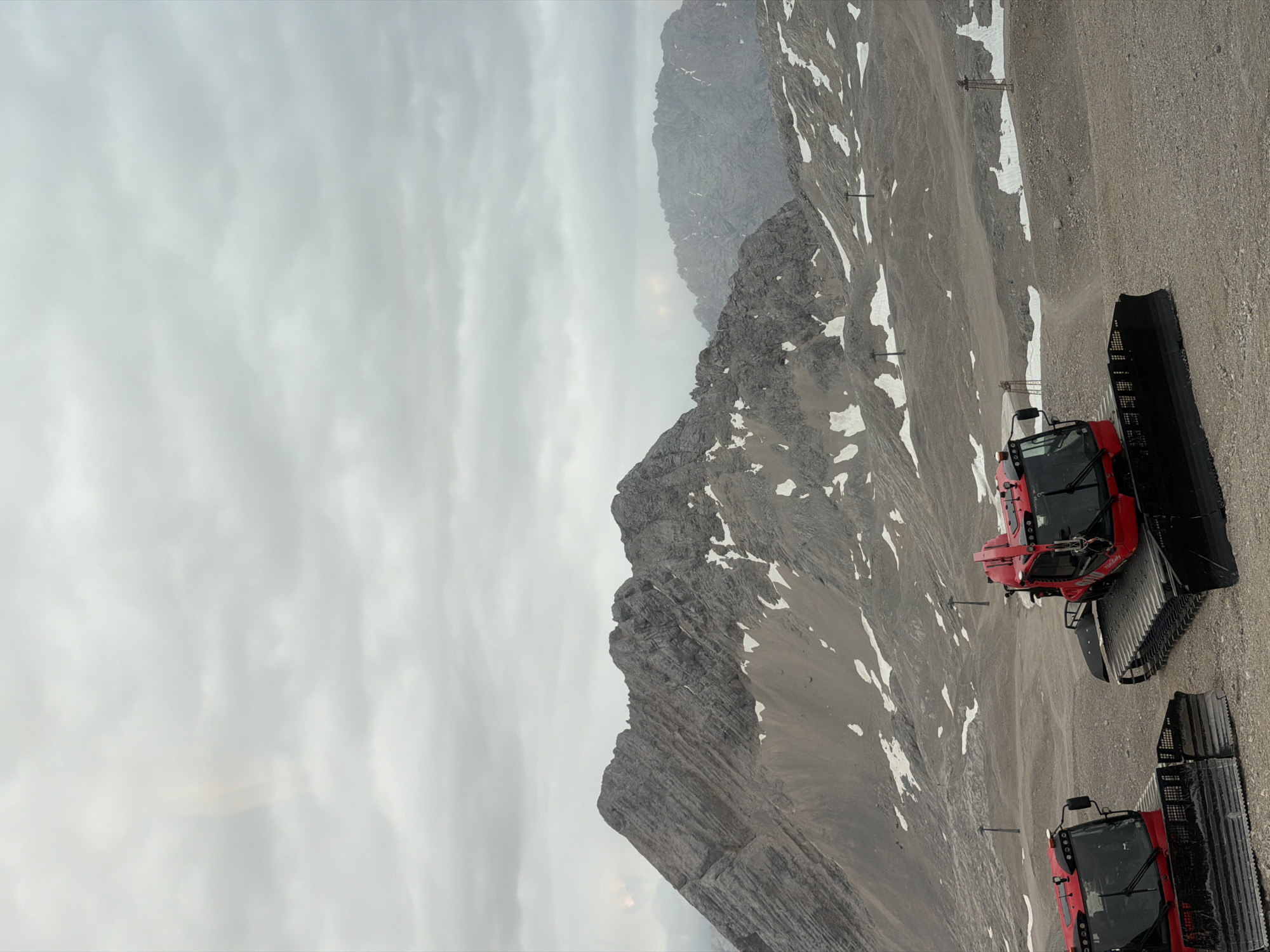 The image shows a mountainous, rocky landscape under a cloudy sky. Two red snow groomers are parked on a gravelly slope. The snow groomers have large, black treads and visible cabins with tinted windows. Ski lift poles are dotted across the terrain, some with small structures at the top, suggesting this is a ski resort or a mountain area that experiences snow. Patches of snow are visible on the mountainsides, even though the ground is mostly rocky and gravelly. The lighting suggests it is daytime, but the overcast sky creates a diffused light. The overall mood is industrial and utilitarian, focused on the machinery and the rugged environment. The location is identified as Wildermieming, Austria. No people are visible in the image.
