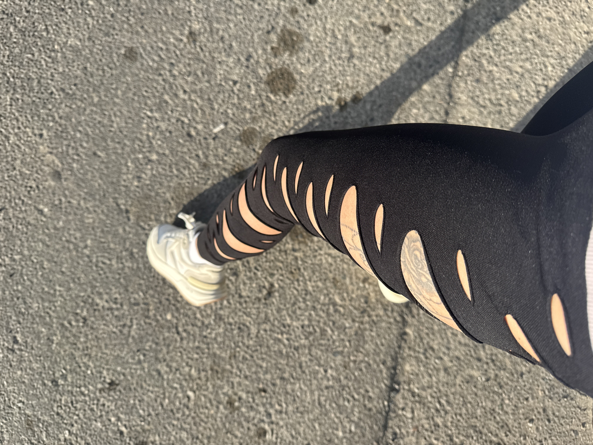 The image depicts a person's leg clad in black leggings with decorative cuts, revealing skin underneath. The leggings appear to be on a female-presenting person. A partially visible white sneaker is present on the left foot. The leg is positioned on a paved surface, likely a sidewalk or street. The texture of the pavement is visible. Shadows are cast on the pavement, indicating a light source from the side. There's no other identifying context for the location, and specifically, there is no contextual information present in the photograph that would allow for the identification of this being in the city of Batumi, Georgia.