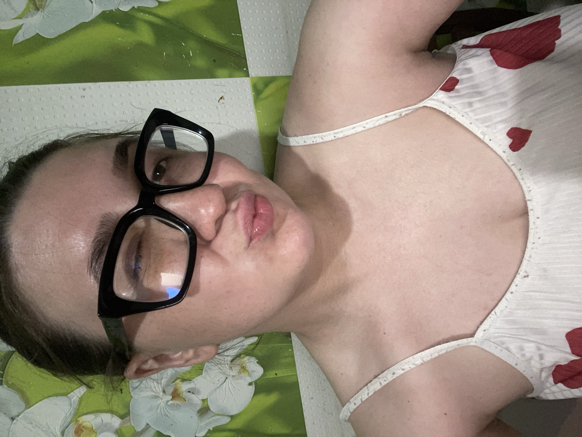 A woman lies on a patterned surface. She wears glasses and a white top with red heart designs. The background is a green and white floral pattern. There is no information available to ascertain the location as Chaikovsky, Russia.