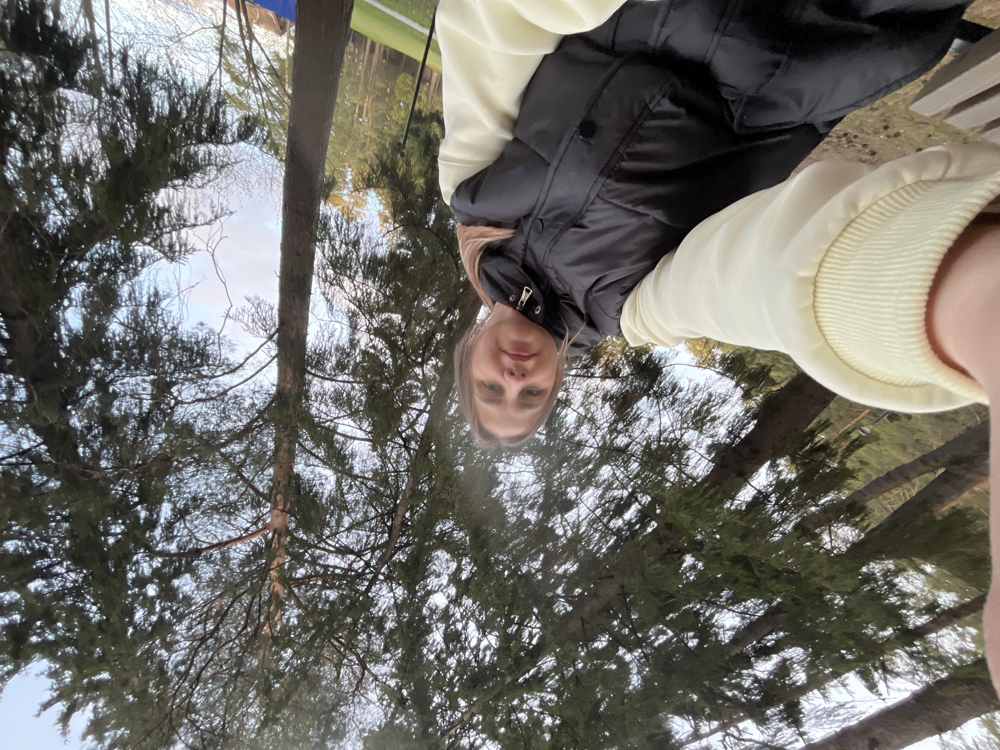 The image displays a vertically oriented outdoor selfie, featuring a young woman looking directly at the camera from a slightly low angle. She has light-colored hair and a neutral expression. She is dressed in a black puffy vest layered over a light-colored, possibly cream or pale yellow, long-sleeved sweater or sweatshirt.
The background is primarily composed of a dense canopy of evergreen trees, characterized by their green needles and visible brown trunks, suggesting a forest or park setting. Patches of overcast sky are visible through the tree branches. In the distant upper right, behind the trees, faint hints of human presence are discernible, including a green area (possibly a field or banner) and a blue structure. A light-colored wooden structure, potentially a bench or railing, is partially visible on the right side, behind the woman's arm.
The scene takes place outdoors during the daytime, likely in a natural environment such as a park or forest in Votkinsk, Russia. The diffused lighting suggests an overcast sky or possibly late afternoon. The woman's attire indicates cool or mild weather conditions. There is no visible text in the image.