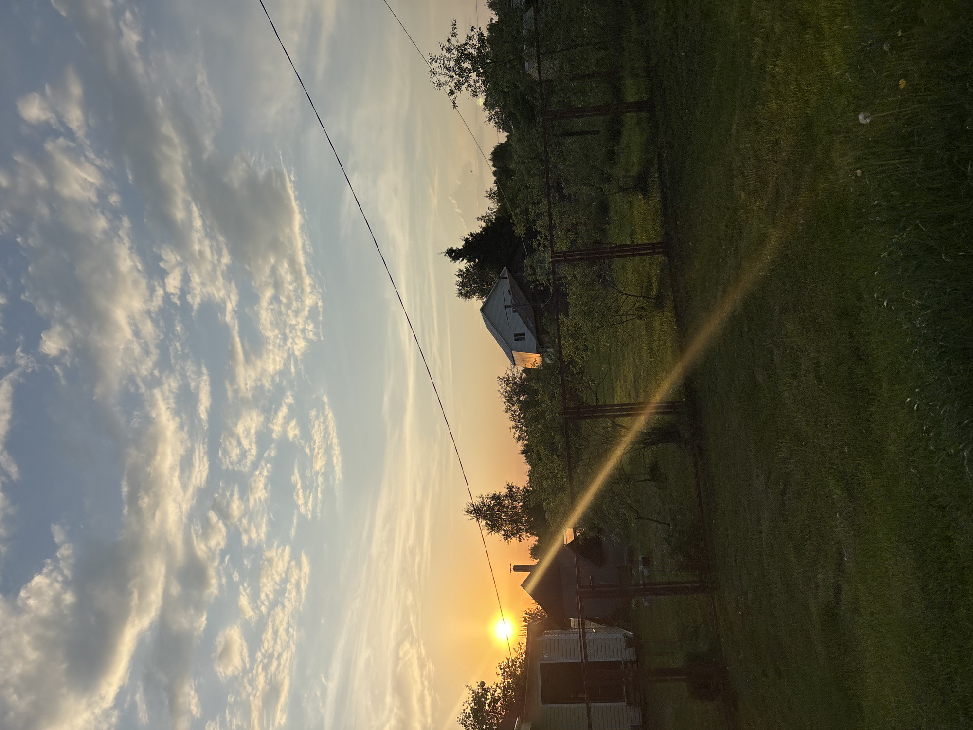 The image shows a rural landscape at sunset. A house is visible in the middle ground, and power lines stretch across the sky. The sky is filled with clouds tinged with orange from the setting sun. Green grass covers the ground, and trees line the horizon. The location is potentially in Мещёры, Russia, based on the query. There are no people visible in the image.