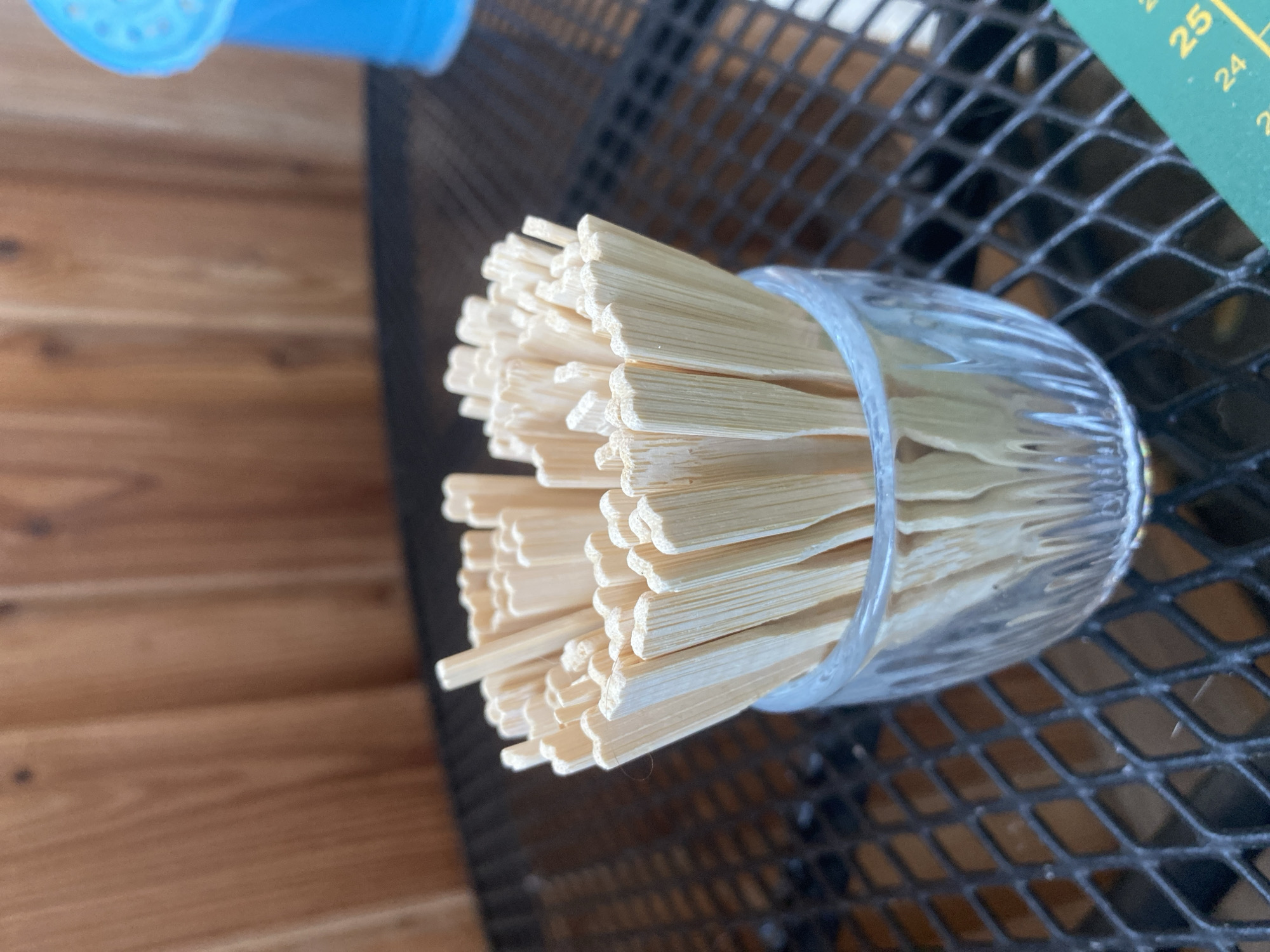 The image shows a close-up of a glass filled with wooden skewers. The skewers are arranged neatly and appear to be unused. The glass is clear with a ribbed texture around its base. The background features a wooden surface to the left, possibly a table or wall, and a dark, metallic mesh surface to the right, which could be part of outdoor furniture. A small portion of a green object with yellow markings, possibly a ruler or measuring tape, is visible in the upper right corner. The markings on the green object include the numbers "25" and "24". The lighting suggests it might be daytime, and the setting appears to be outdoors or in a well-lit indoor space. There is no indication of people or any specific activity taking place. The focus is entirely on the skewers and the glass container.
