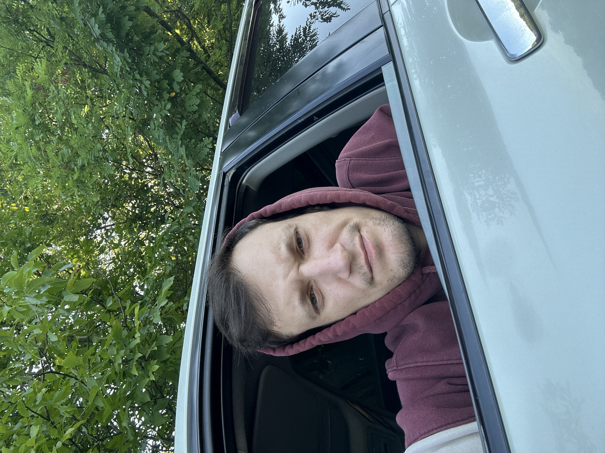 A man is photographed from the chest up, leaning out of the open window of a car. He is wearing a maroon hooded sweatshirt with the hood pulled up. His expression is neutral, with his eyebrows slightly furrowed. The background consists of green trees and foliage, suggesting an outdoor, possibly rural or park-like setting. The car appears to be light-colored, possibly white or silver, and the interior is visible through the window, including a dark dashboard. The image captures a close-up view of the man's face and upper body. There is no indication of the man's specific location in Makiv, Ukraine; the image only shows him in a car by some trees.