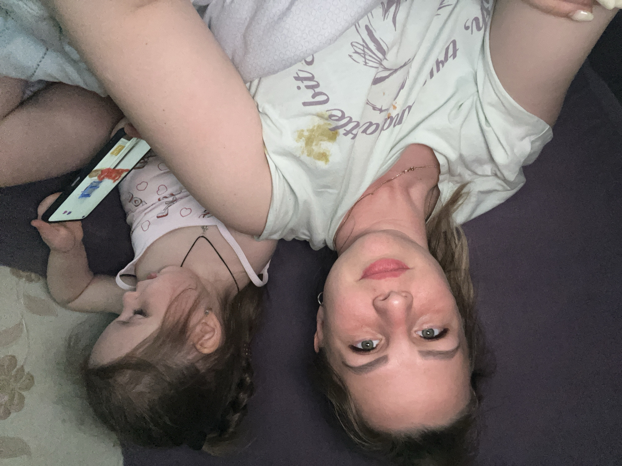 A woman and a young child lie head-to-head on a dark purple surface, possibly a bed or sofa. The woman is fair-skinned with green eyes and wears a light green t-shirt with stains on the front. She has a thin gold necklace. The child, who has fair skin and brown hair in braids, wears a white tank top with heart patterns. The child is holding a smartphone and is looking at the screen. The woman is looking directly at the camera. A patterned cushion is visible to the left of the child. The setting is most likely an indoor domestic environment. There is no indication of Izhevsk, Russia.