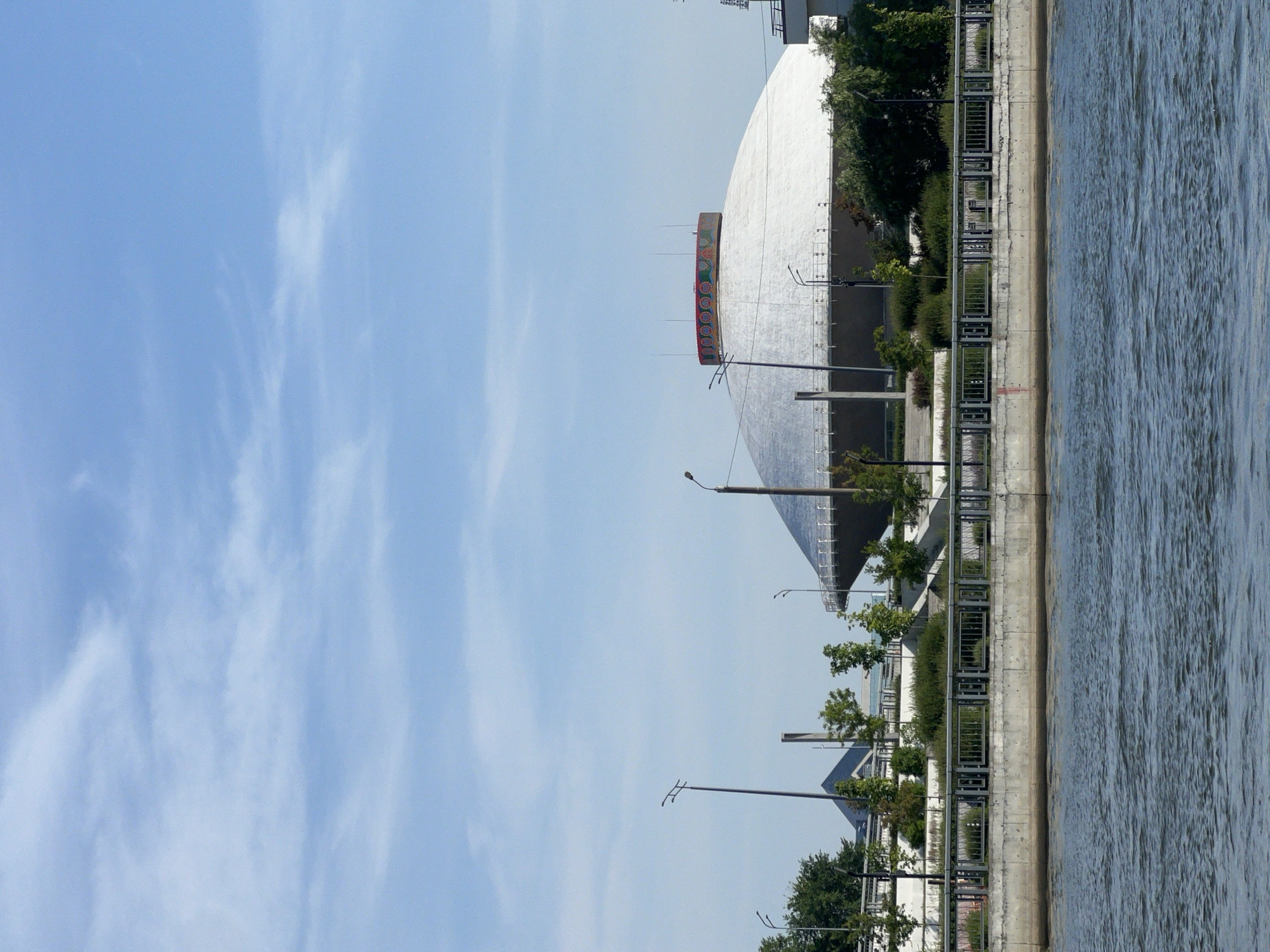This is a daylight, eye-level, medium shot of a large, white, domed building with a decorative, circular band around its top, situated on the bank of a wide river in Kazan, Russia. The building appears to be the Kazan Circus. The sky is blue with scattered white clouds. In the foreground, the choppy blue water of the river fills the bottom half of the frame. Behind the water, a concrete embankment with a metal railing forms a barrier along the river's edge. Beyond the railing, the domed building is visible, along with some trees, bushes, and streetlights. To the left of the dome, two triangular structures are partially visible. The overall scene depicts a sunny day in an urban waterfront environment.