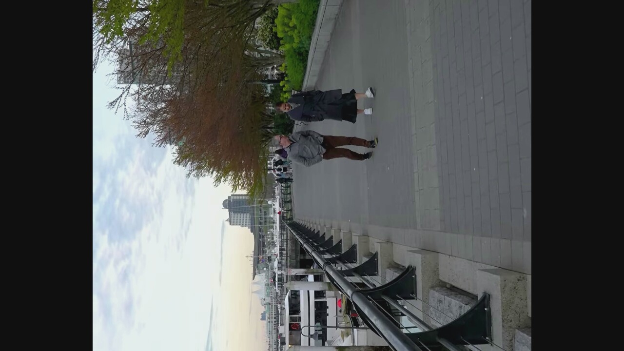The image depicts a waterfront promenade in Vancouver, Canada. Two individuals, a man and a woman, are walking side-by-side on the paved walkway. The man, on the left, wears a grey jacket and brown trousers. The woman, on the right, is dressed in a dark trench coat and a long skirt. Behind them, a line of trees with sparse, autumnal foliage borders the walkway. In the background, several boats are docked along the waterfront, and beyond that, the cityscape of Vancouver rises, featuring modern buildings and structures. The sky above is overcast with scattered clouds. The scene captures a moment of casual stroll along the waterfront, with the urban environment and natural elements creating a characteristic Vancouver vista.
