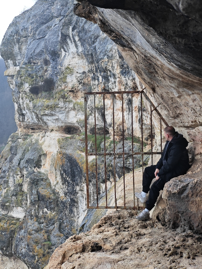 The image displays an adult male seated within a natural rock formation on a cliff face in the Shumen Plateau Nature Park, Shumen, Bulgaria. The man is positioned towards the right side of the frame, seen from a three-quarter rear-right view. He wears a dark puffer jacket over a grey hooded garment, dark track pants, and light-colored athletic shoes. He is seated on an uneven, light brown rock surface, leaning against the darker rock wall of a large overhang. His head is turned to the left, and his gaze is directed forward and slightly downward, observing the vista beyond.
A rusty, weathered metal gate, constructed from vertical and horizontal square bars, occupies the central left portion of the frame. This gate stands vertically, seemingly spanning an opening in the rock structure, and is secured to the right rock wall by a diagonal metal support bar. Beyond the gate and to the left, a large, mottled grey and beige limestone cliff face with visible layers, cracks, and patches of green moss and lichen descends steeply. The environment is rugged and natural, illuminated by diffused light, indicating an overcast sky.