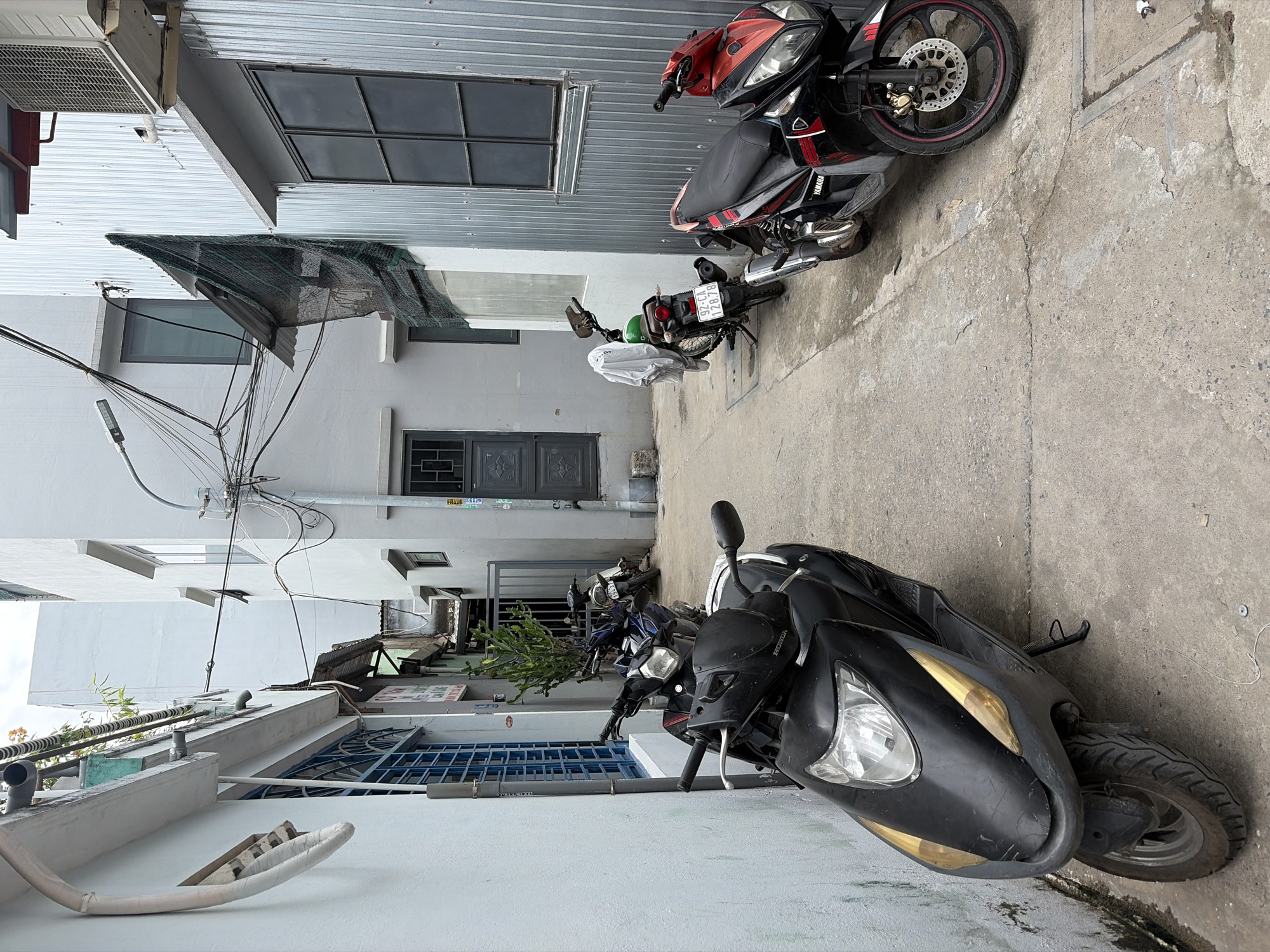 The image depicts a narrow alleyway in Đà Nẵng, Vietnam. On the left, a pale blue wall with a blue-grated window runs alongside the alley. Several motorcycles are parked in the alley. In the foreground, a black scooter is positioned on the left, facing forward. Further down the alley, a dark blue motorcycle is parked. To the right, a green motorcycle with a white cloth draped over its handlebars is parked, and behind it, a black and red Yamaha motorcycle is facing the viewer. The alley is paved with concrete, and the buildings on either side are of varying materials, including corrugated metal and light gray concrete. Overhead, a dense network of electrical wires crisscrosses the alley, supported by poles and attached to the buildings. The sky is overcast.