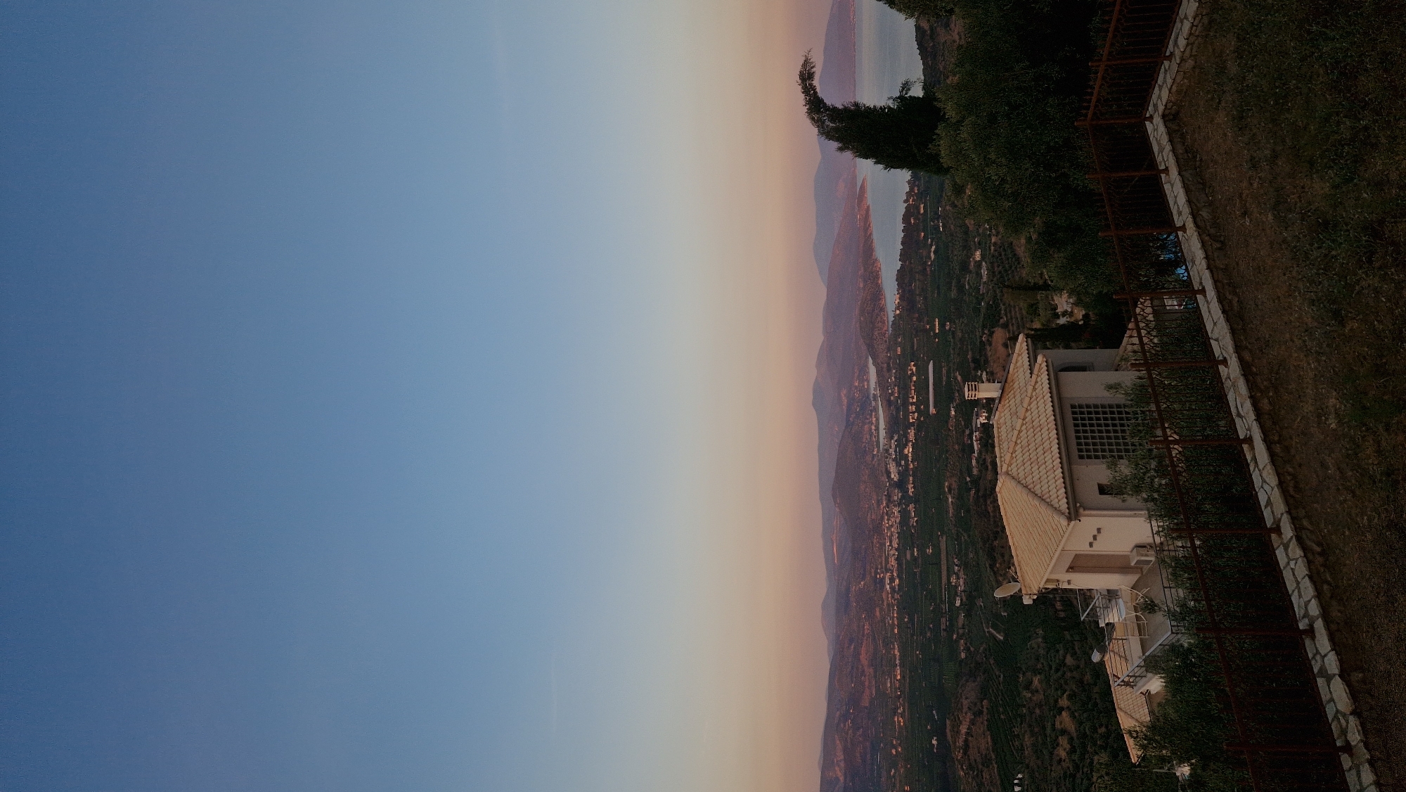 An aerial view of the Municipal Unit of Asini, Greece, at dusk showcases a villa on a hilltop overlooking a coastal landscape. The sky transitions from a clear, light blue at the top to a gradient of soft pink and orange hues near the horizon. Below the sky, a panoramic vista unfolds, featuring rolling hills covered in olive groves and scattered buildings of a town nestled in the valley. The sea is visible in the distance, meeting the rugged coastline and distant mountains.
In the foreground, a light-colored villa with a tiled roof is prominently situated on the left side of the frame. It features a terrace with outdoor seating and a distinctive section of white gridded windows. A dark metal fence encloses the property, separating it from a grassy area in the immediate foreground. A few trees, including a strikingly curved dark green tree on the right, are interspersed within the landscape, adding depth and texture. The overall scene depicts a serene, picturesque evening in a Mediterranean setting.