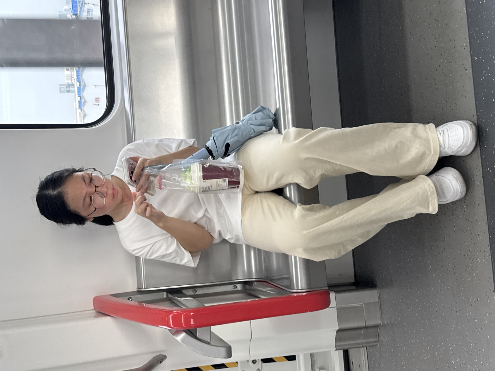 A young woman with dark hair, wearing glasses, a white t-shirt, and light-colored pants, sits on a train seat in Shunde District, China. She is holding a cell phone in her right hand and a clear plastic bag containing a beverage and an umbrella in her left. The umbrella, light blue in color, is folded and rests beside her on the seat. The train interior is visible, with metallic seats and a red handle on the left side of the frame. A window behind her shows an urban landscape with buildings and what appears to be a body of water. The floor of the train car is dark grey with small white specks.