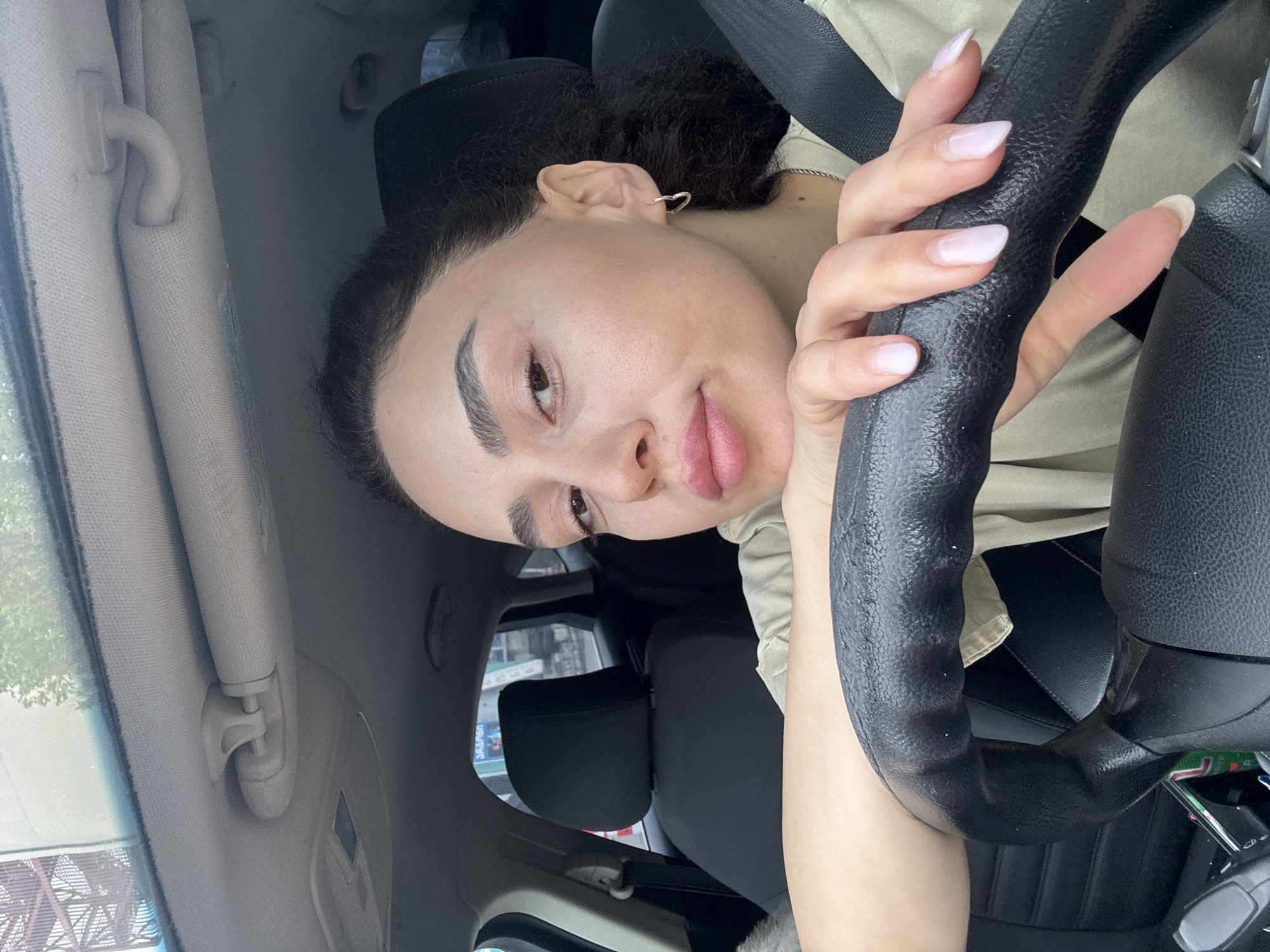 A light-skinned woman with dark curly hair is in the driver's seat of a car. She is wearing a tan or light green shirt and a seatbelt. She is holding the black steering wheel with manicured nails. The interior of the car is visible, with a light-colored sun visor and a black headrest. Outside the car window, buildings are visible, suggesting an urban environment. The location appears to be Tbilisi, Georgia, as evidenced by the presence of Georgian language signage visible in the background.