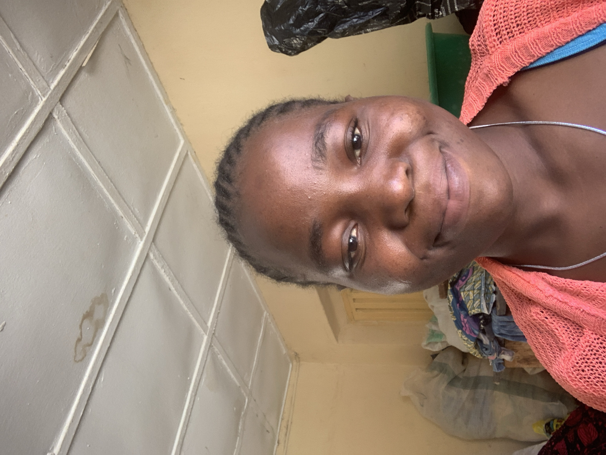 The image depicts a woman with dark skin and braided hair. She is wearing a pink, mesh-like garment over a blue top, and a silver necklace. The background includes a wall with a patterned, rectangular design, painted white. The wall has a light brown stain. There is also a pile of clothes and plastic bags in the background, alongside a green object. The location is specified as Jos, Nigeria, but there is no visual evidence within the image to confirm or deny this location.