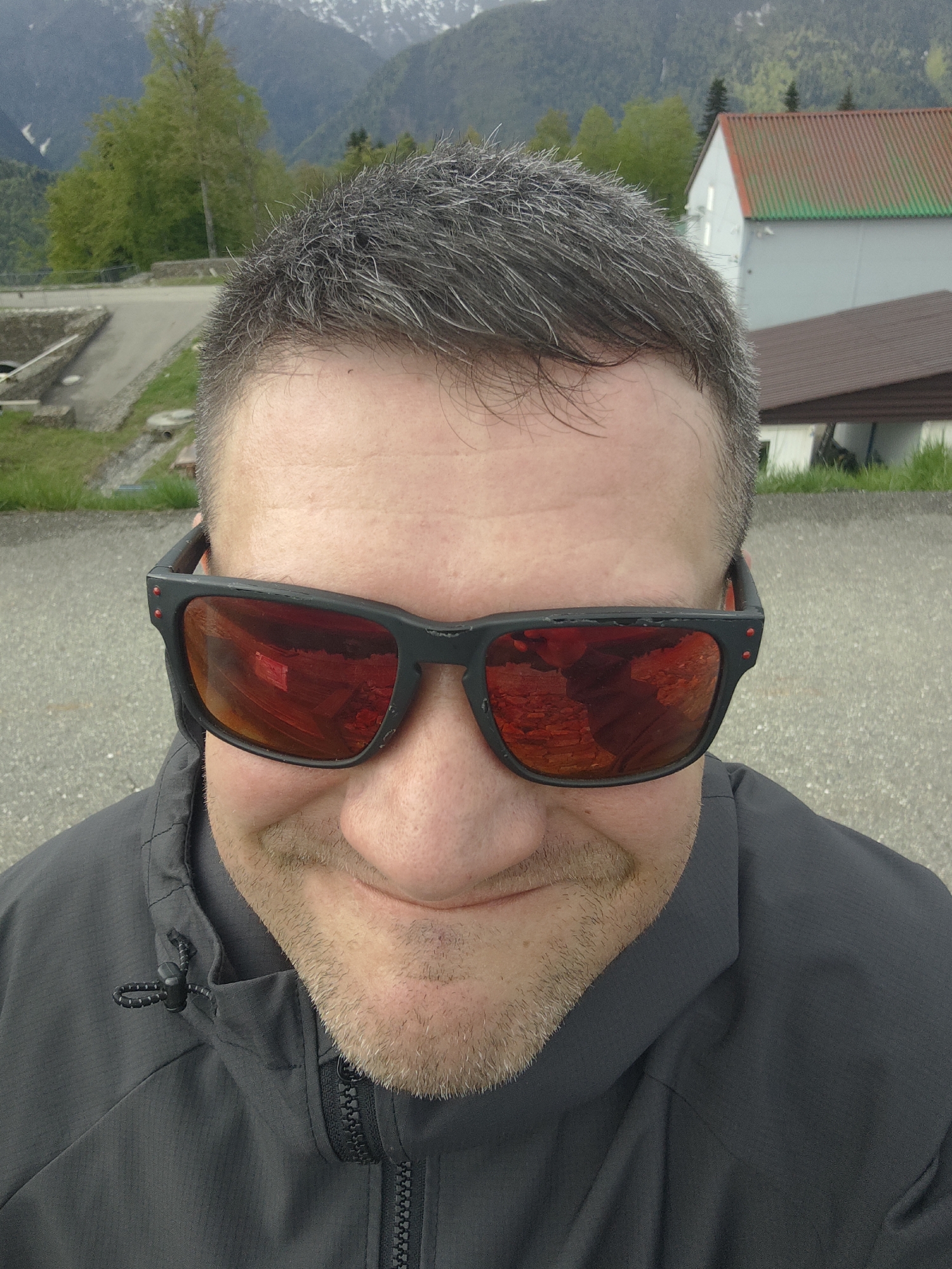 A close-up shot from a high angle shows a middle-aged man with graying hair and a light beard. He wears black sunglasses with red lenses that reflect a building. He has on a gray jacket. In the background are trees, a building with a red and gray roof, and snow-capped mountains. The location is the city of Unknown, Russia.