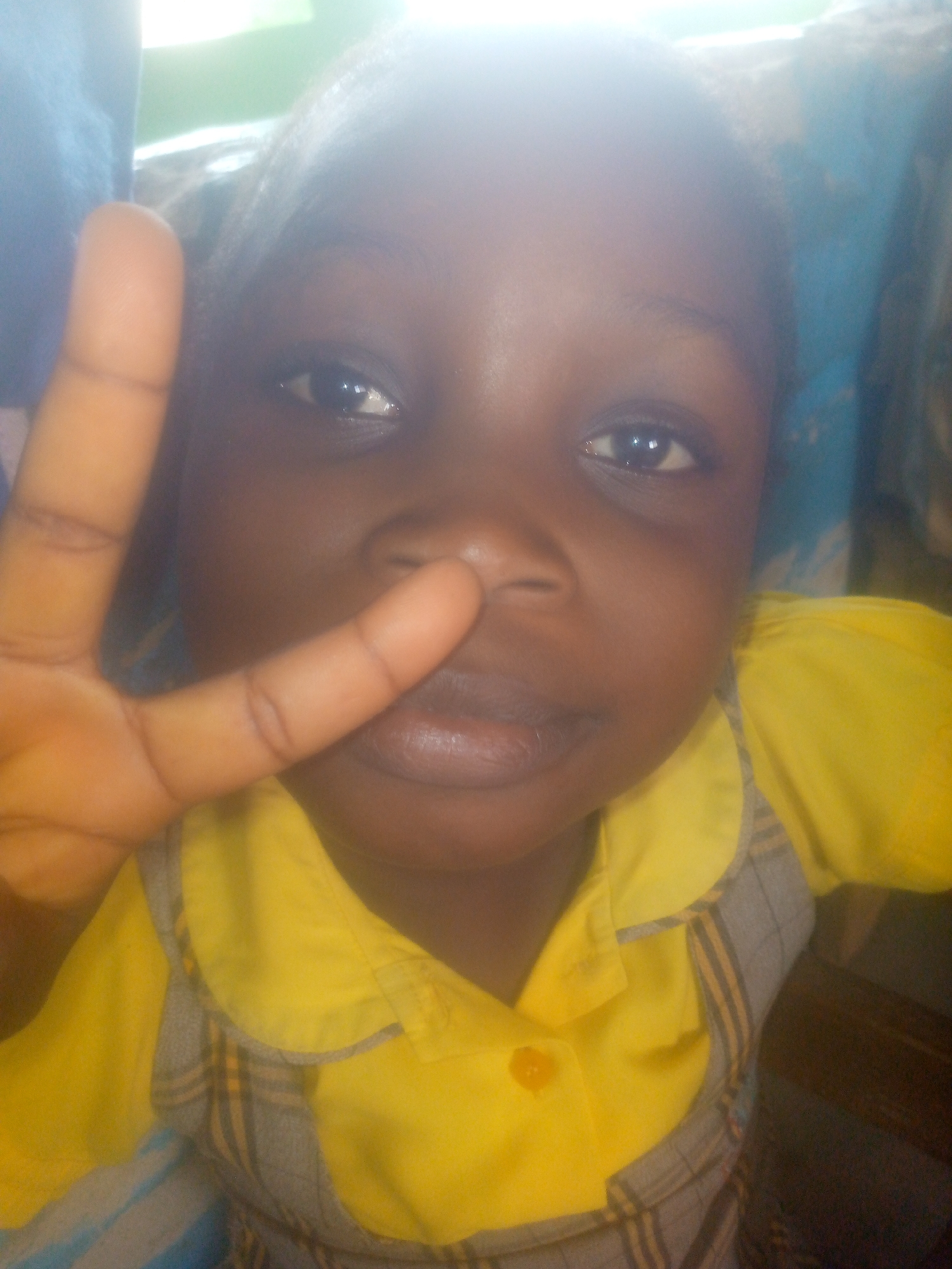 A close-up shot of a child with dark skin, wearing a yellow shirt with a gray and yellow plaid pattern around the collar. The child is holding their fingers in a peace sign, with the fingers positioned near their nose. The background is blurred, but appears to include blue and green elements. The location of the scene is Unknown, Unknown.