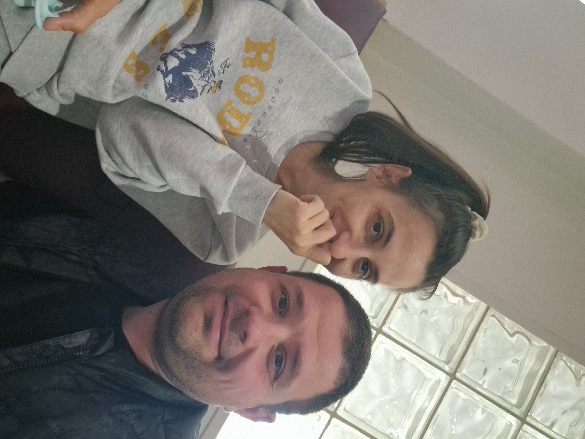 A slightly tilted, medium close-up portrait shows two individuals, a man and a woman, indoors in Bearpark, United Kingdom. The man, positioned in the foreground, has short dark hair and stubble, and is wearing a dark jacket or top. He looks directly at the camera with a neutral to slightly smiling expression.
Behind and to his right, the woman is seen wearing a light grey sweatshirt. The sweatshirt features a prominent yellow text 'ROD' with smaller text 'MOUNTAIN' beneath it, and the word 'Let's' next to a blue graphic resembling a mountain lion or similar animal. She has dark hair, partially tied back, and wears a thin silver necklace. Her right hand is casually positioned near her mouth as she looks towards the camera with a calm gaze.
The background consists of a plain light-colored wall or ceiling and a distinct section of translucent glass block windows, suggesting an indoor setting with diffused light. The overall scene conveys a casual and personal moment, likely a self-taken photograph.