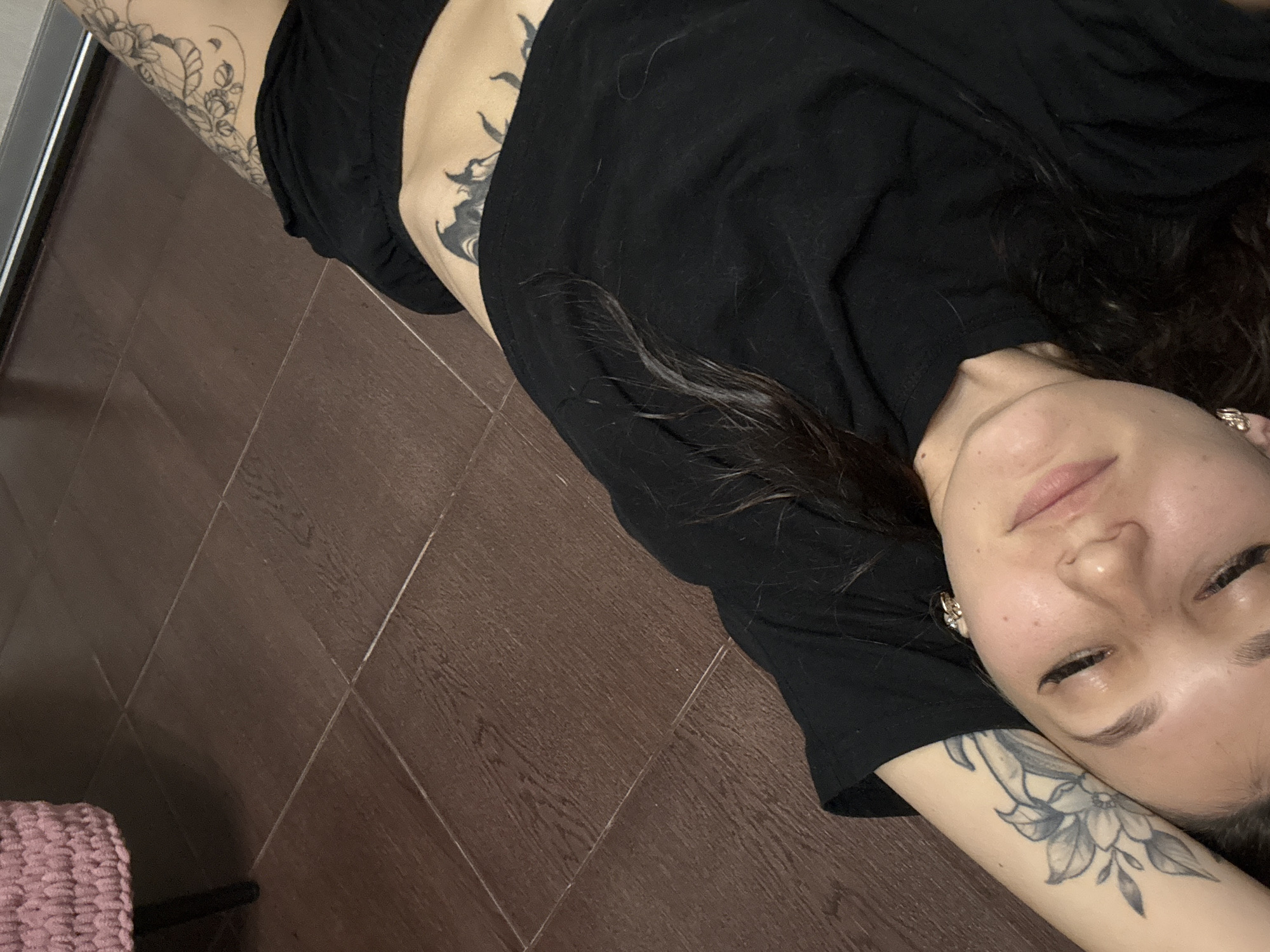 A young woman with dark hair is lying on her back on a tiled floor. She is wearing a black t-shirt and black shorts. She has tattoos on her left arm and on her lower abdomen. The room appears to be a bedroom, with a pink knitted blanket visible in the upper left corner. The lighting is dim. The location is not specified in the image.
