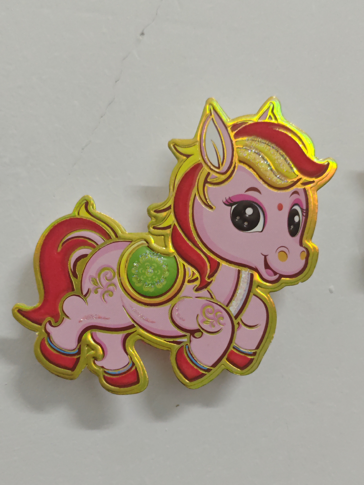 A shiny, cartoonish pony sticker is adhered to a white, slightly textured surface, possibly a wall. The pony is pink with red and yellow hair, a green saddle, and gold trim. The image was captured in a setting consistent with Chongqing High-tech Zone, China, though no specific location details are discernible from the image alone. There are no people visible.