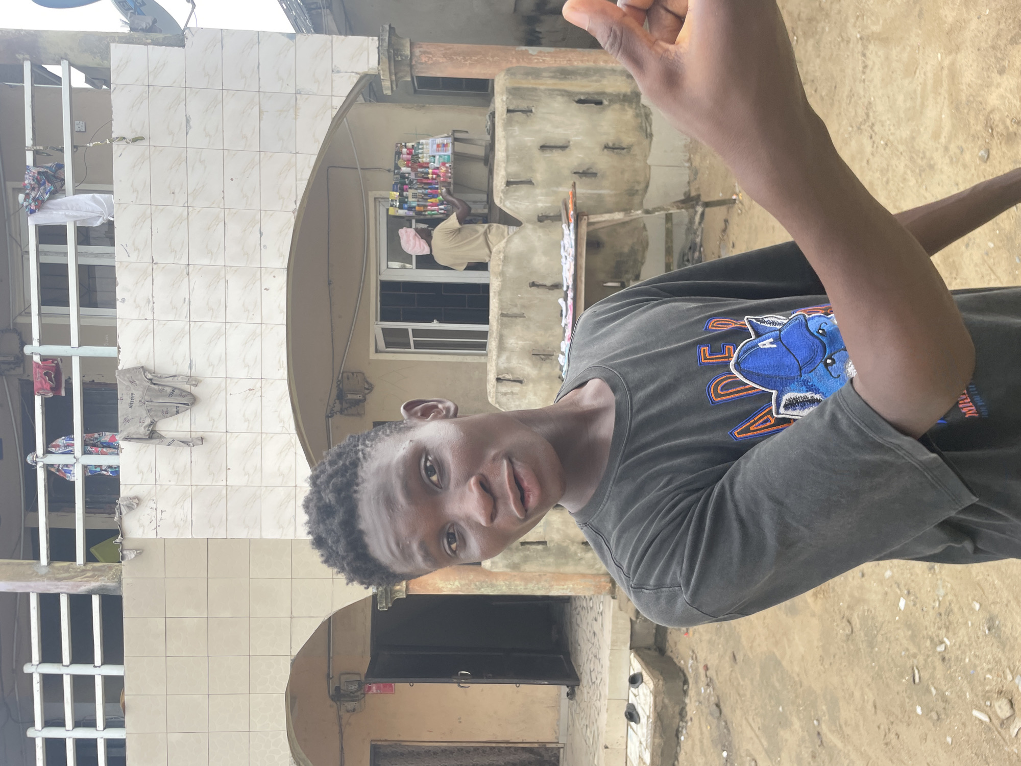 The image, taken in Badagry, Nigeria, is rotated 90 degrees clockwise, showing a young Black man in the foreground looking directly at the camera. He wears a dark grey t-shirt featuring a prominent blue and orange graphic of an animal's head, possibly a wolf or dog, with partially visible letters like "U" and "S" on either side. His right arm is raised, and part of his left arm is also visible.
Behind him, a multi-story building serves as the backdrop. Its facade is a mix of light-colored, patterned tiles and rough concrete sections. On a balcony or elevated platform of this building, a person wearing a light-colored top and a pink head covering stands behind a display stall. The stall is laden with numerous small, colorful bottles, likely indicating a vendor selling cosmetics, perfumes, or similar items. Below this vendor, a window with white frames and dark grates is visible.
To the left of the main subject, portions of white metal railings suggest balconies or window grates, with some clothes, including a light grey long-sleeved shirt and colorful fabric, hanging to dry. The ground in the foreground is unpaved and appears to be dirt or sand. Further in the background, a satellite dish is mounted on the building's roof. The scene is illuminated by natural daylight, suggesting it is daytime. No strong shadows are visible. The overall setting depicts a bustling urban or semi-urban environment.