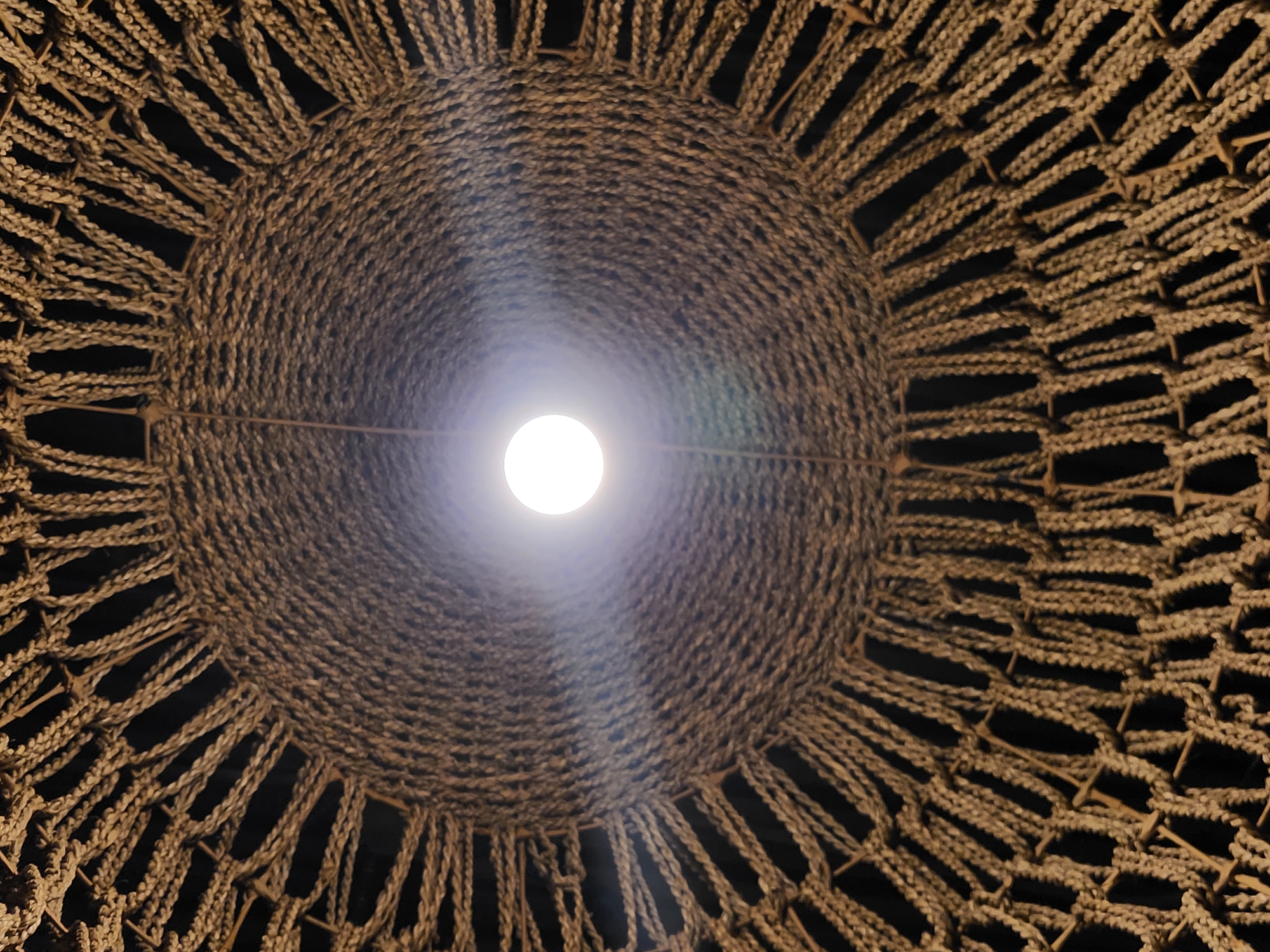 The image shows a close-up, low-angle view of a woven lampshade made of natural fibers, possibly jute or sisal. The lampshade has a circular opening in the center, from which a bright, white light bulb is illuminated. The woven texture creates a radial pattern, with strands of rope tightly packed towards the center and becoming more spaced out towards the edges. The background is dark, suggesting the lampshade is hanging indoors, possibly in a dimly lit area. There are no people or specific landmarks visible. The image does not provide any information about its location in Maitland, United States of America.
