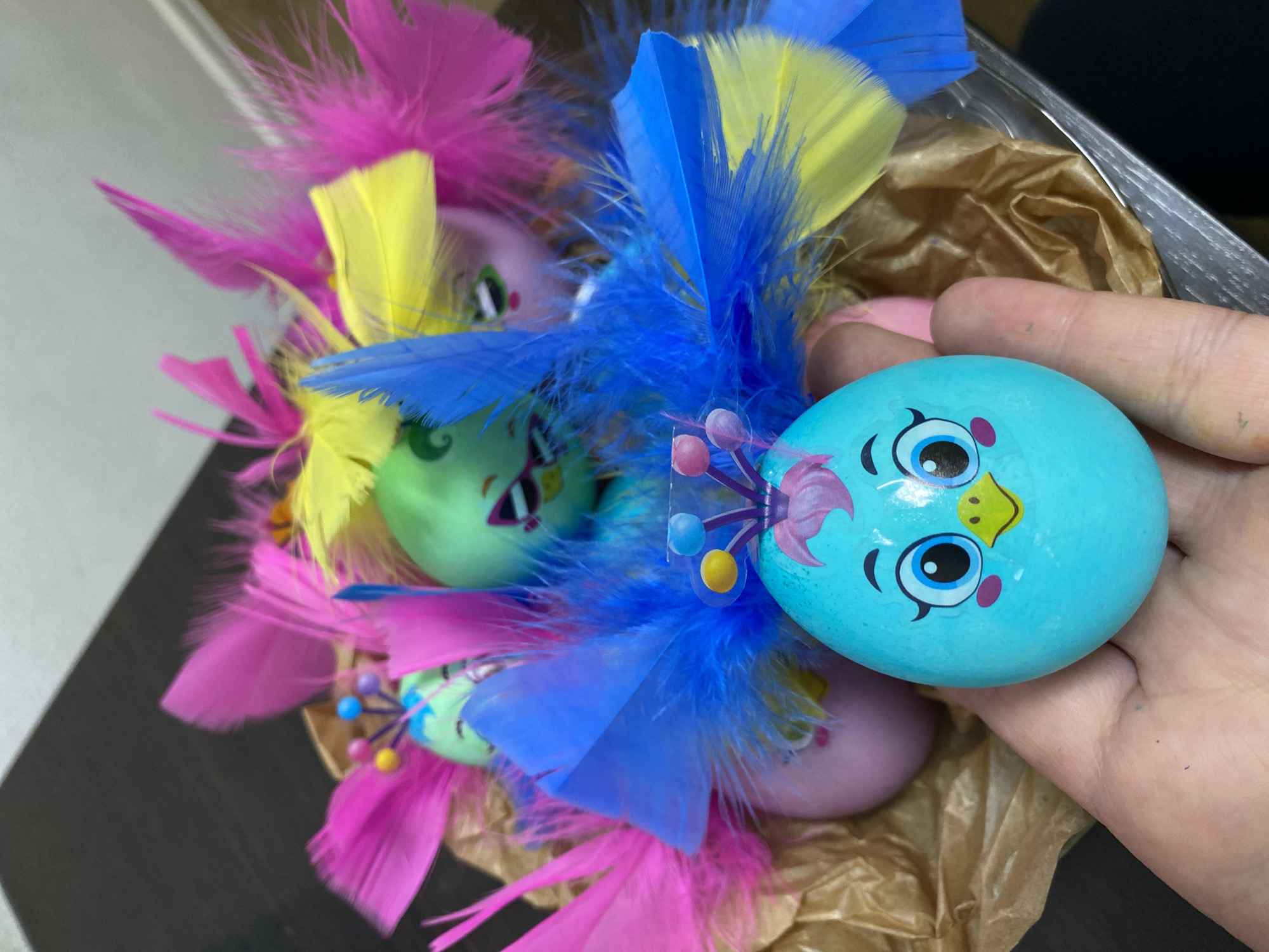 A hand holds a blue Easter egg decorated with a cartoon bird face. The egg is part of a larger arrangement of other decorated Easter eggs nestled among brightly colored feathers (pink, yellow, blue). The eggs rest on a bed of crinkled brown paper inside a clear plastic container. The setting is inside. There is no indication of it being in the city of Chaikovsky, Russia.
