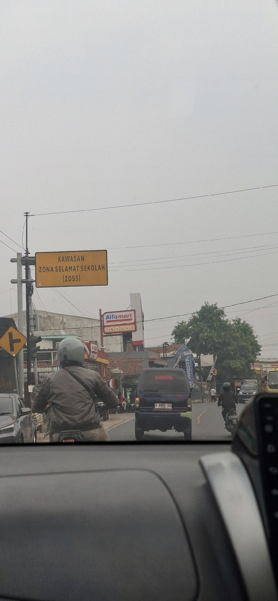 The image captures a street scene in Sirnagalih, Indonesia, likely during daylight hours given the overcast sky. In the foreground, the interior of a vehicle is visible, suggesting the photo was taken from inside a car. The perspective looks forward, showing the road ahead.
Dominating the scene is a bright yellow traffic sign with black lettering that reads "KAWASAN ZONA SELAMAT SEKOLAH (ZOSS)". This translates to "School Safety Zone". The sign is mounted on a pole to the left of the road.
On the road, a person is riding a motorcycle, wearing a grey jacket and a helmet. They are positioned in the center lane, moving away from the camera. To the right of the motorcycle, a dark-colored van is also traveling in the same direction. The van has a license plate visible that reads "D 1003 GD". Further down the road, other vehicles and motorcycles are present, indicating moderate traffic.
On the left side of the road, there is a building with an "Alfamart" sign, which is a convenience store chain. The architecture of the buildings appears to be typical of a commercial area in Indonesia. The environment is somewhat urban, with power lines crisscrossing overhead. A large tree with dense foliage is visible in the background, adding a touch of nature to the otherwise built environment. The overall atmosphere suggests a typical day in a busy Indonesian town or city.