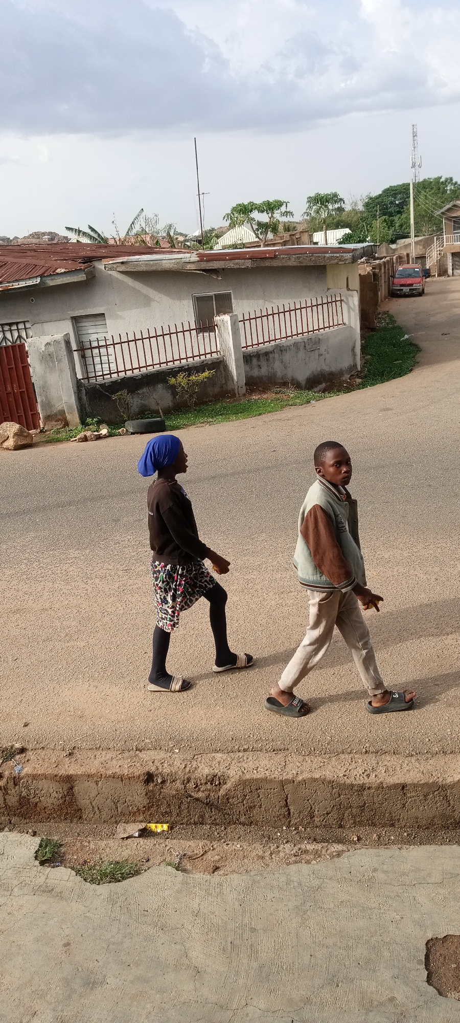 Two children walk side-by-side on a paved road in an urban area of Unknown, Unknown. The child on the left wears a blue head covering, a dark long-sleeved shirt, a patterned skirt, and dark leggings, with sandals on their feet. The child on the right wears a brown and denim jacket, light-colored pants, and sandals. A sidewalk is visible in the foreground, with a road stretching into the background. Several buildings with fences are visible, along with a red vehicle parked in the distance. A utility pole stands among the buildings. The sky is overcast with white and gray clouds.