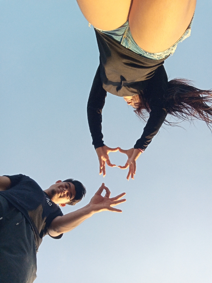 A low-angle shot captures a unique perspective of two individuals against a clear sky. The top figure, appearing inverted, wears denim shorts and a black long-sleeved top, forming the upper half of a heart with their hands. The lower figure, a man, gestures similarly, as if to complete the heart. Both figures are angled upwards, creating a dynamic and playful composition that emphasizes unity and connection.